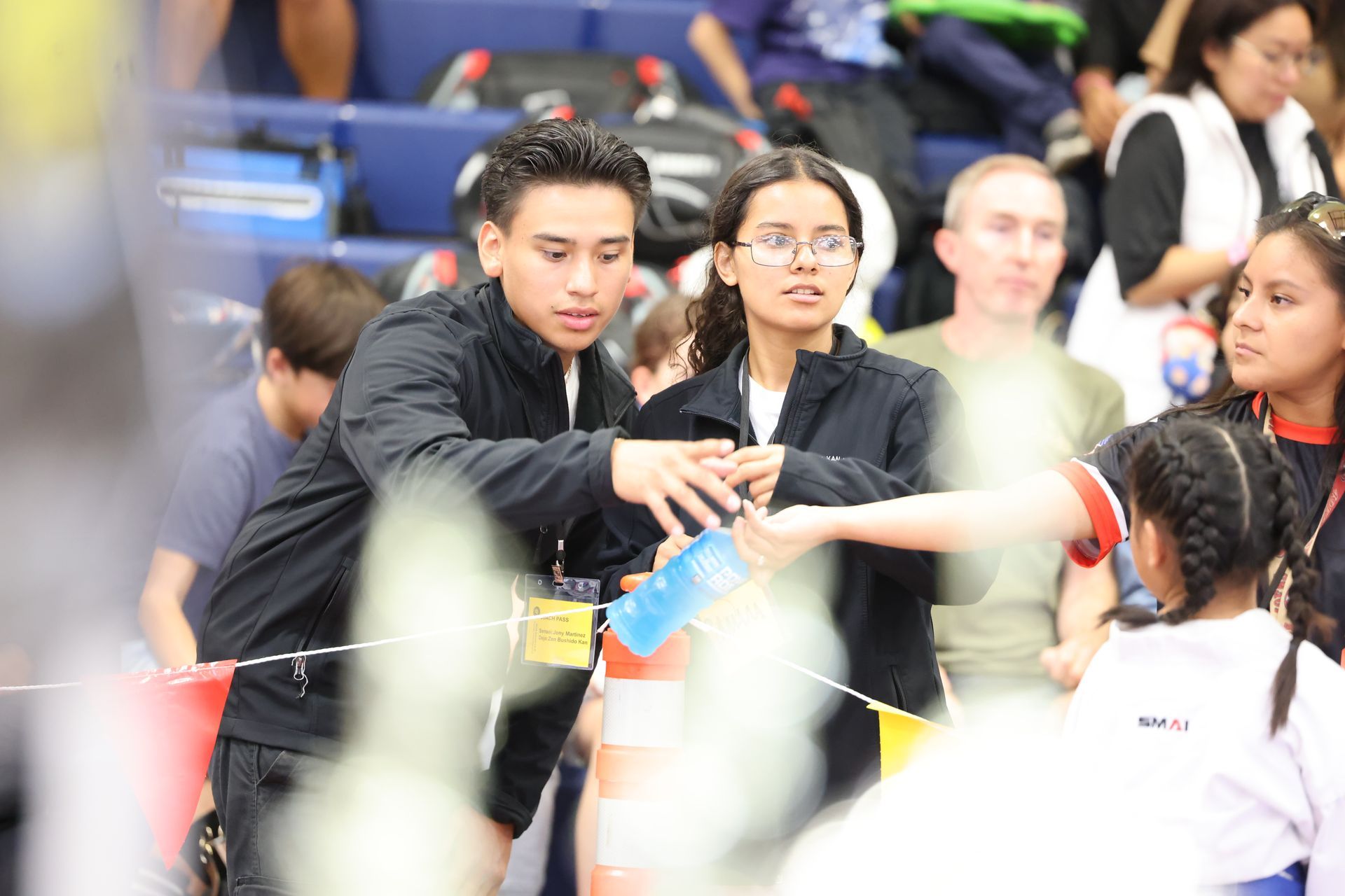 Two people in black jackets, possibly coaches, assist a child at an event. Indoor setting, bright.