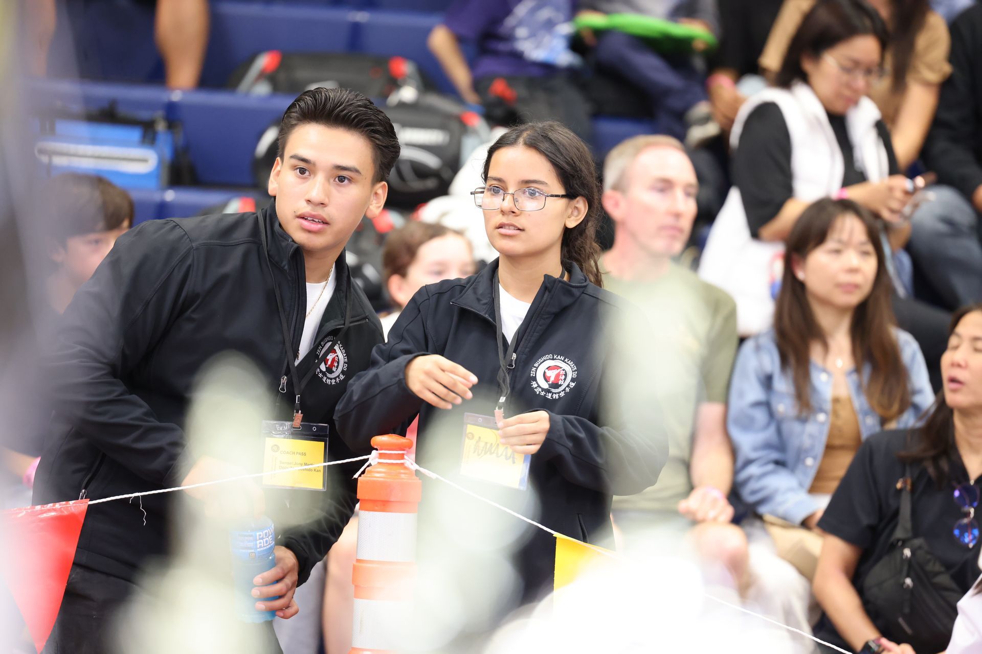 Two young adults with team jackets in a crowd, looking left.