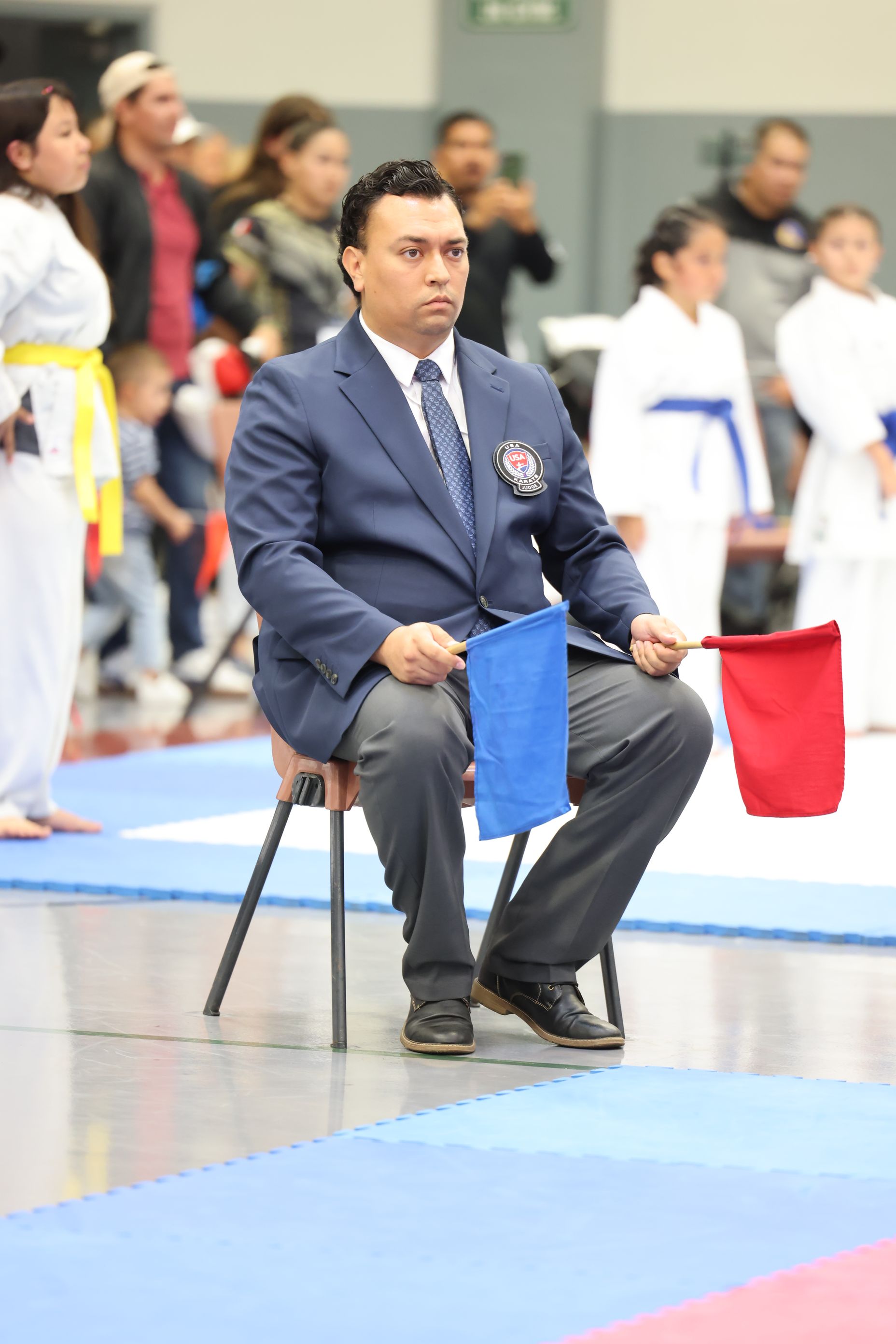 Karate judge in a suit, seated, holding red and blue flags; competition in a gym.