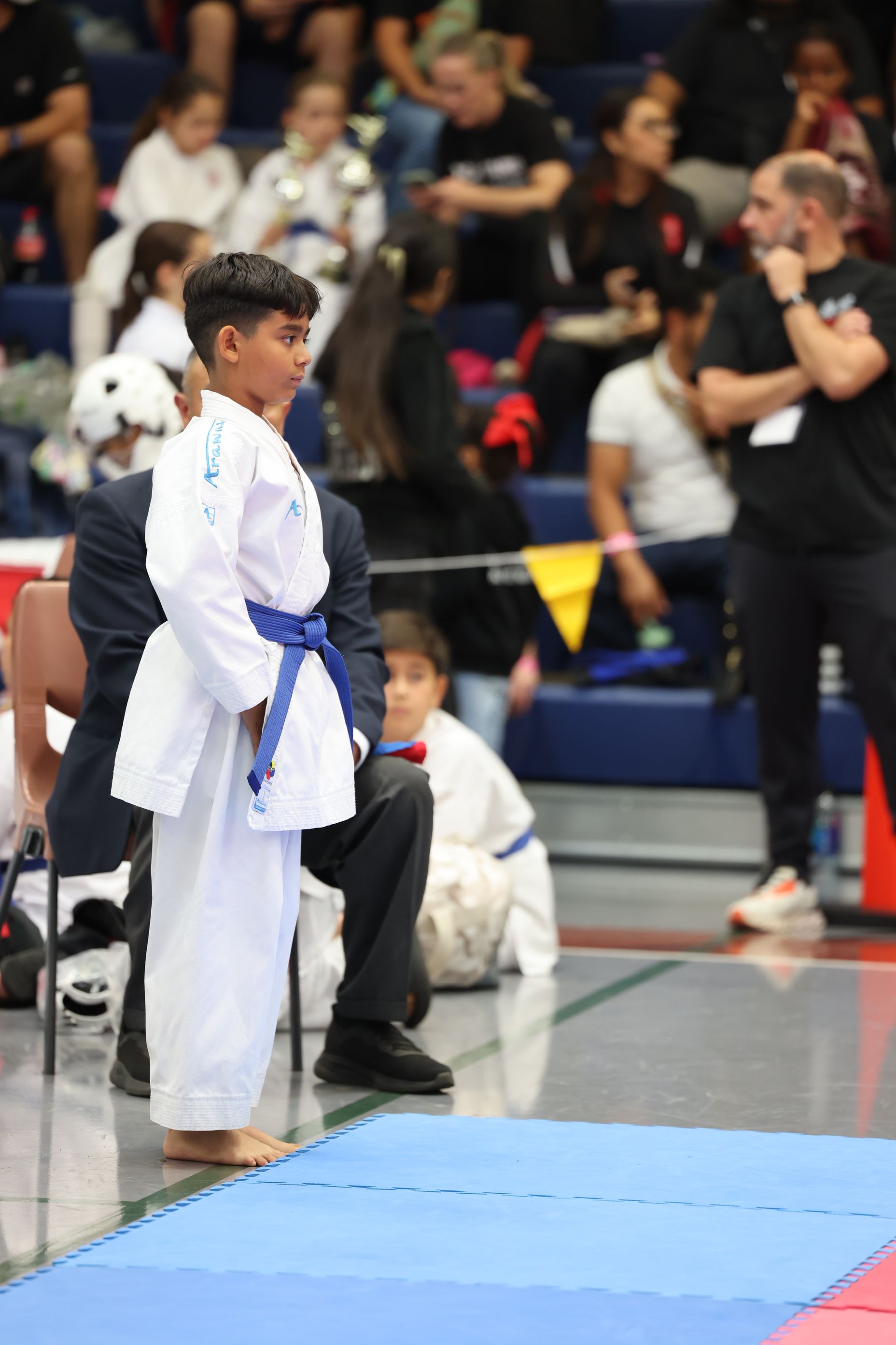 Boy in a white karate gi with a blue belt stands at a competition, spectators in background.