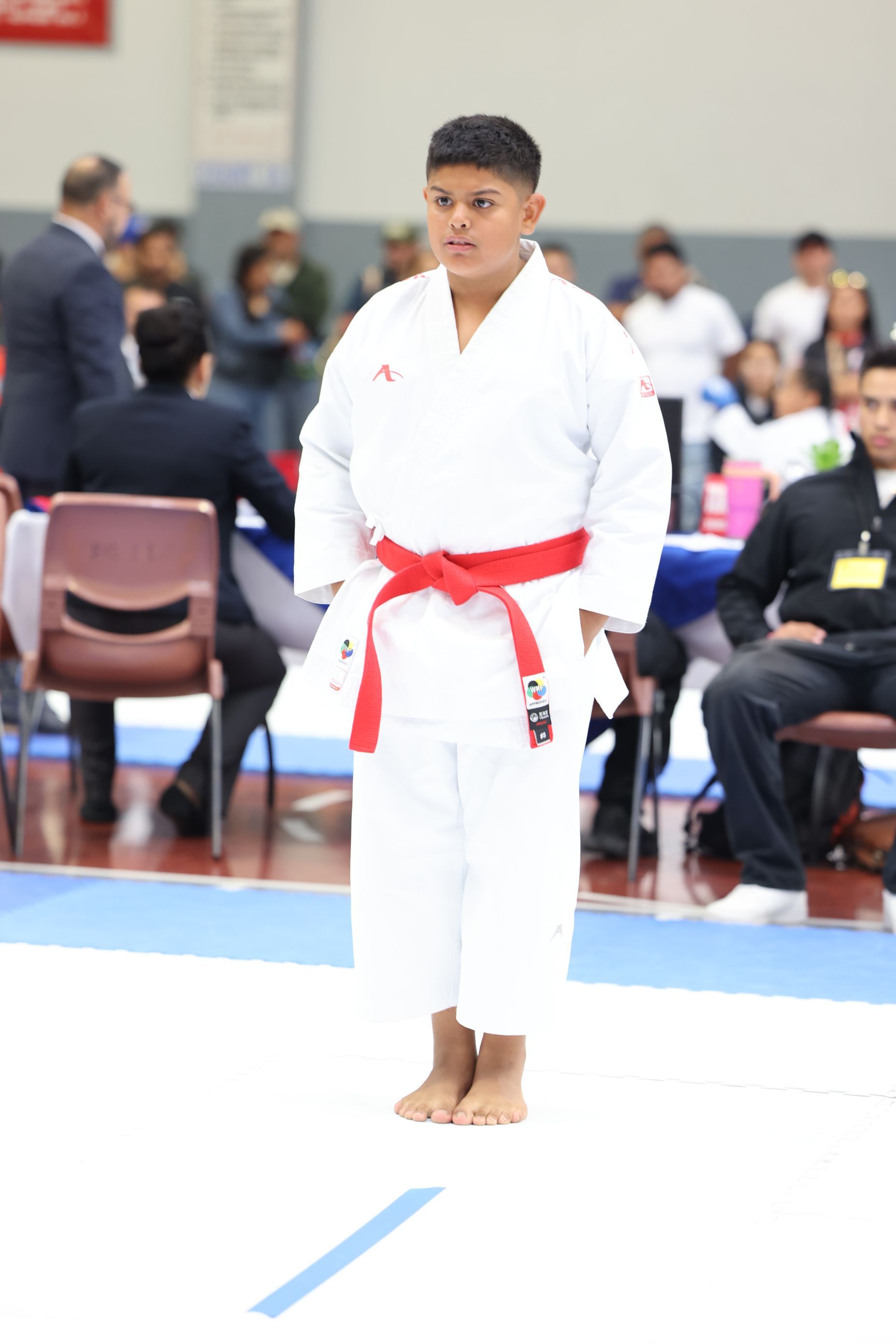 Young karateka in white gi and red belt, standing in a competition setting, hands at sides.