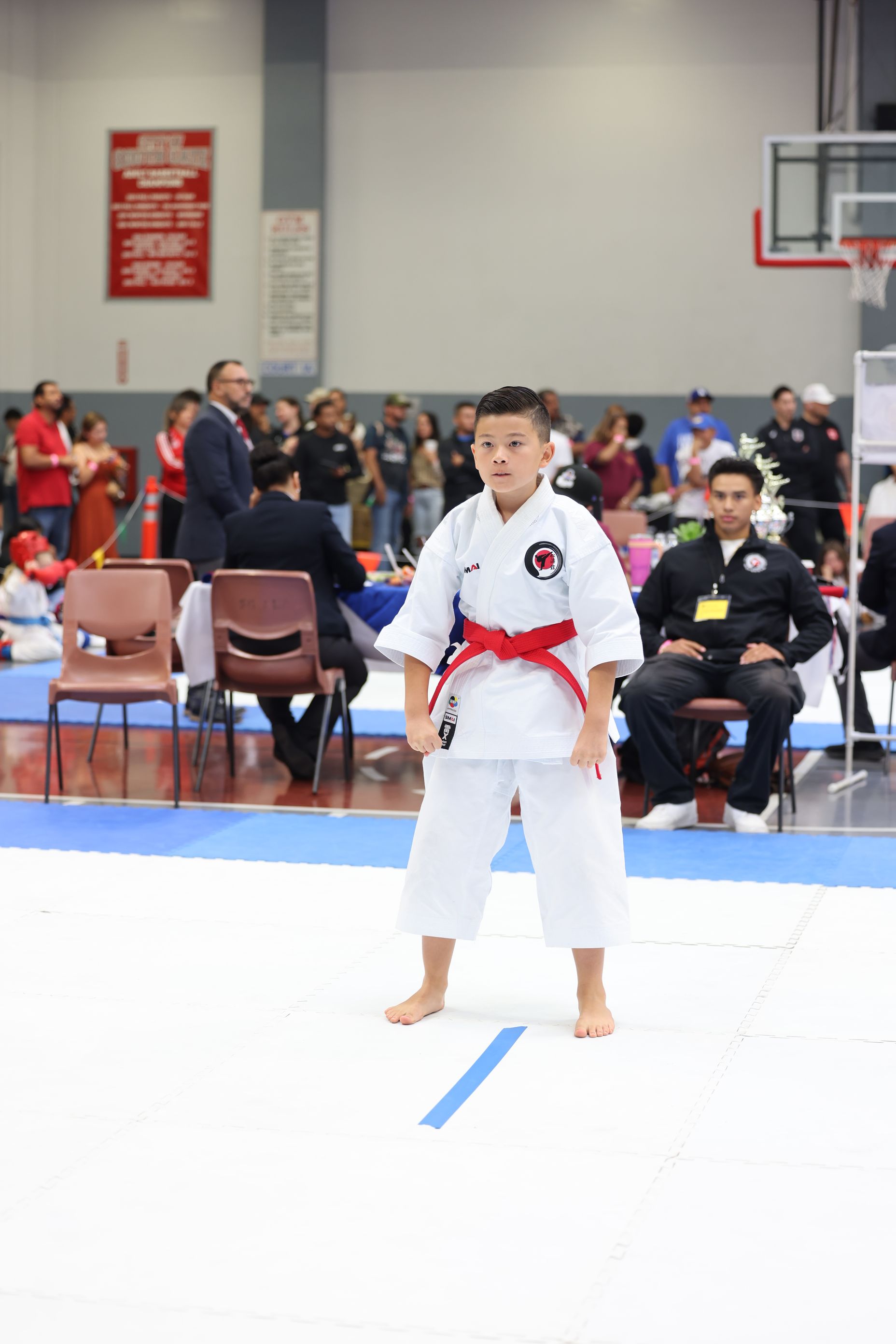 Young karateka in white uniform with red belt stands on mat, indoor competition setting.