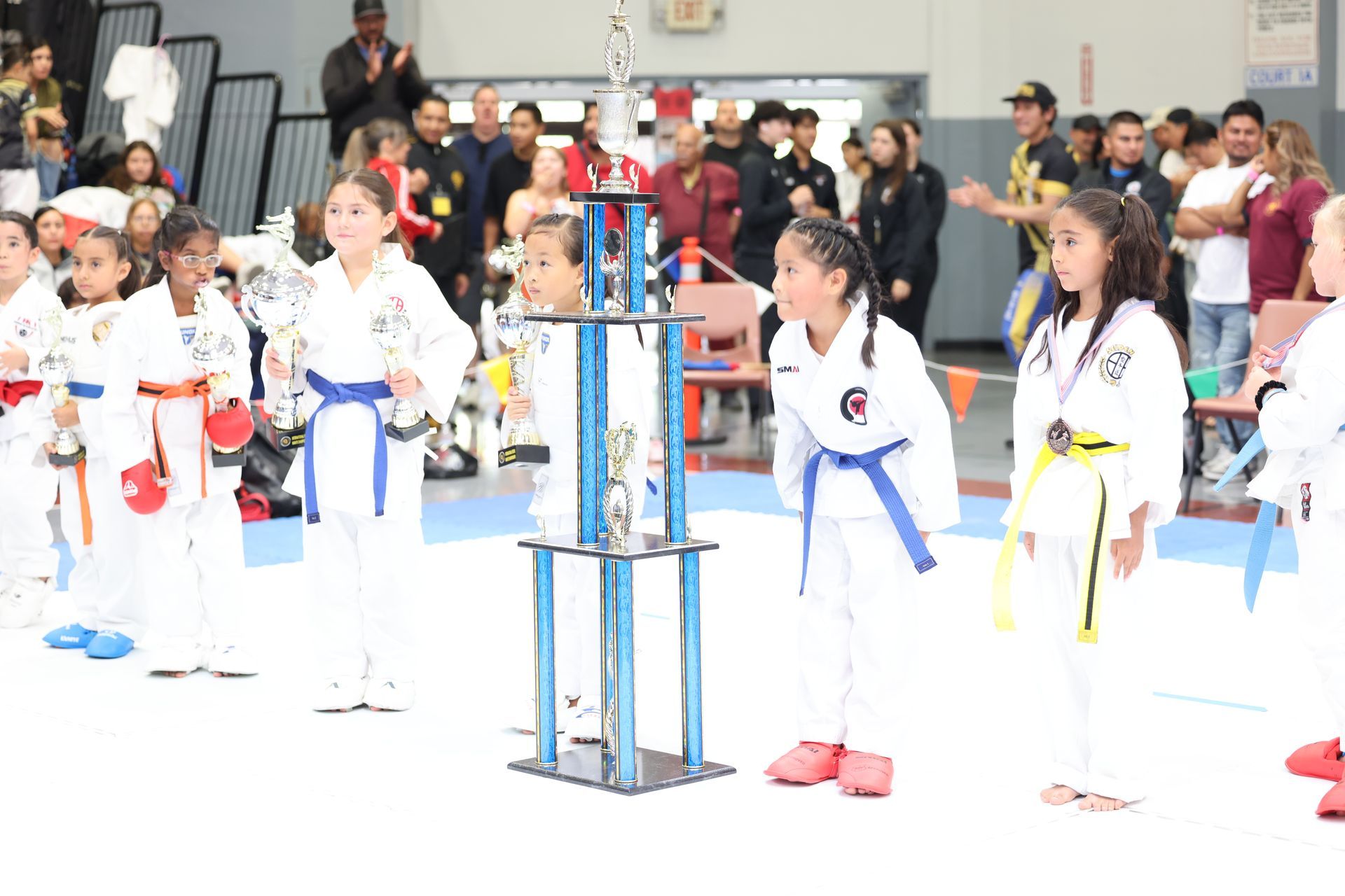Young karate students stand near a trophy. Some wear belts and medals, smiling. An indoor competition.