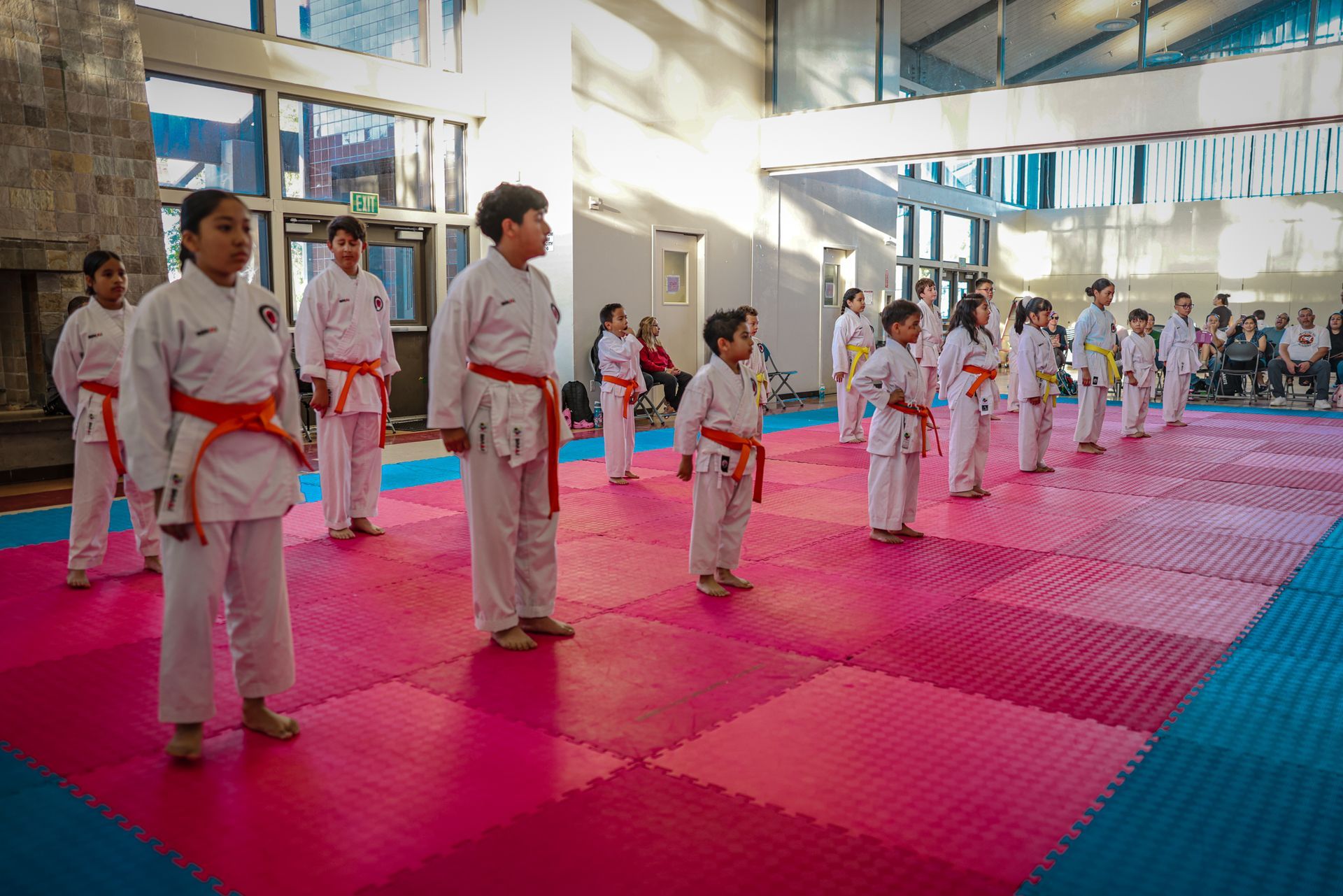 A row of children in white karate uniforms with colorful belts practicing on a pink and blue matted indoor floor.