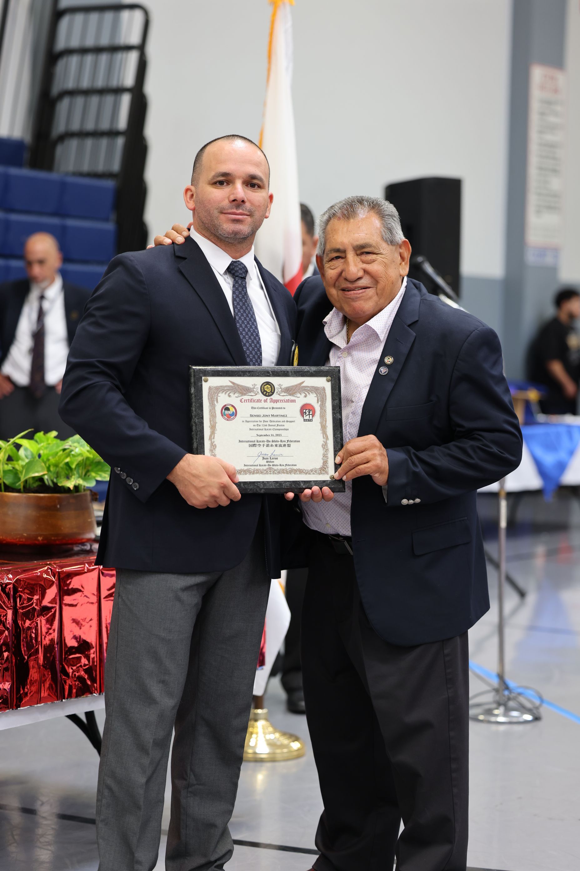 Two men in suits holding a framed certificate; indoors, gymnasium setting.