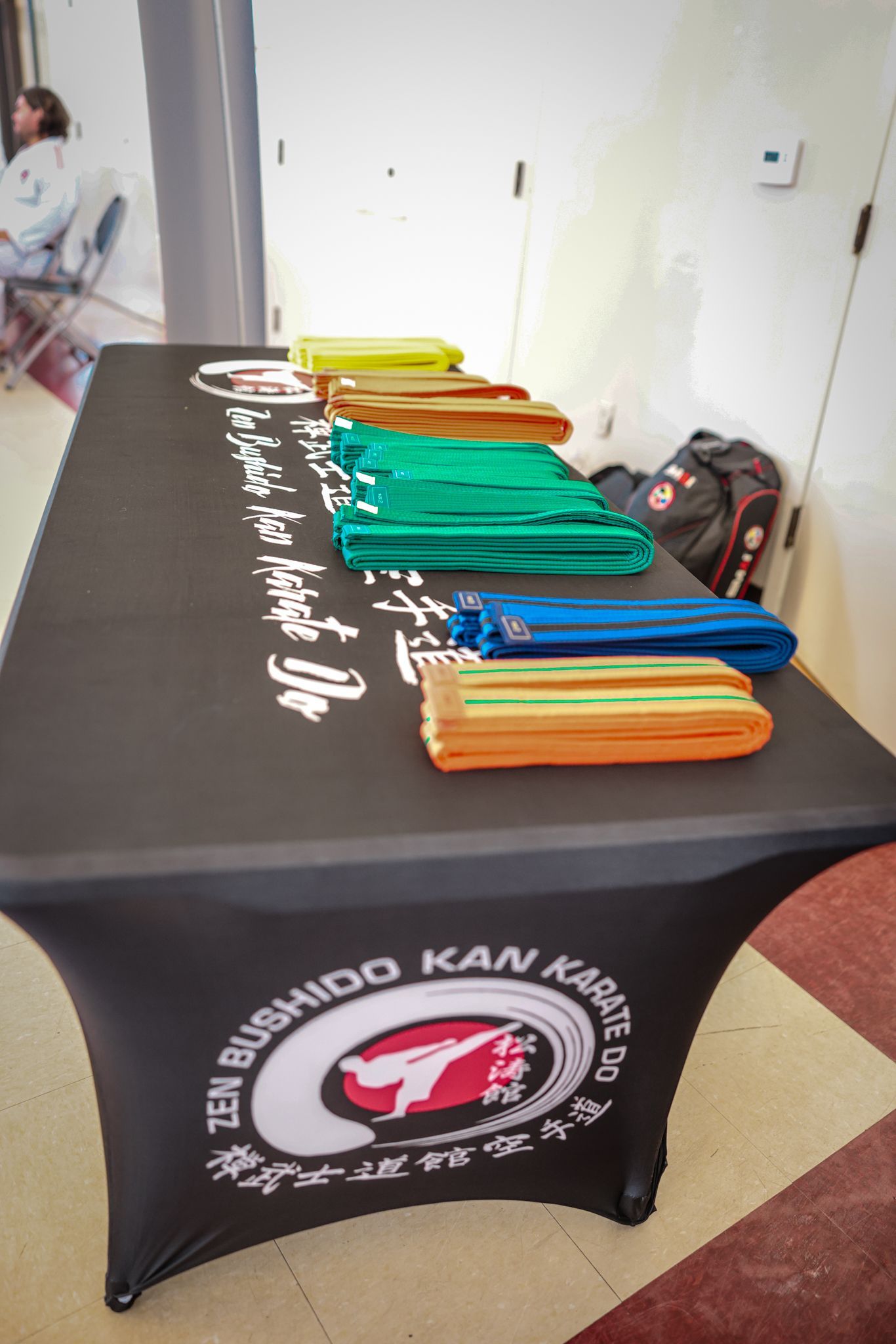 A table covered in a black cloth with a karate logo, holding several folded colored belts, in a studio setting.