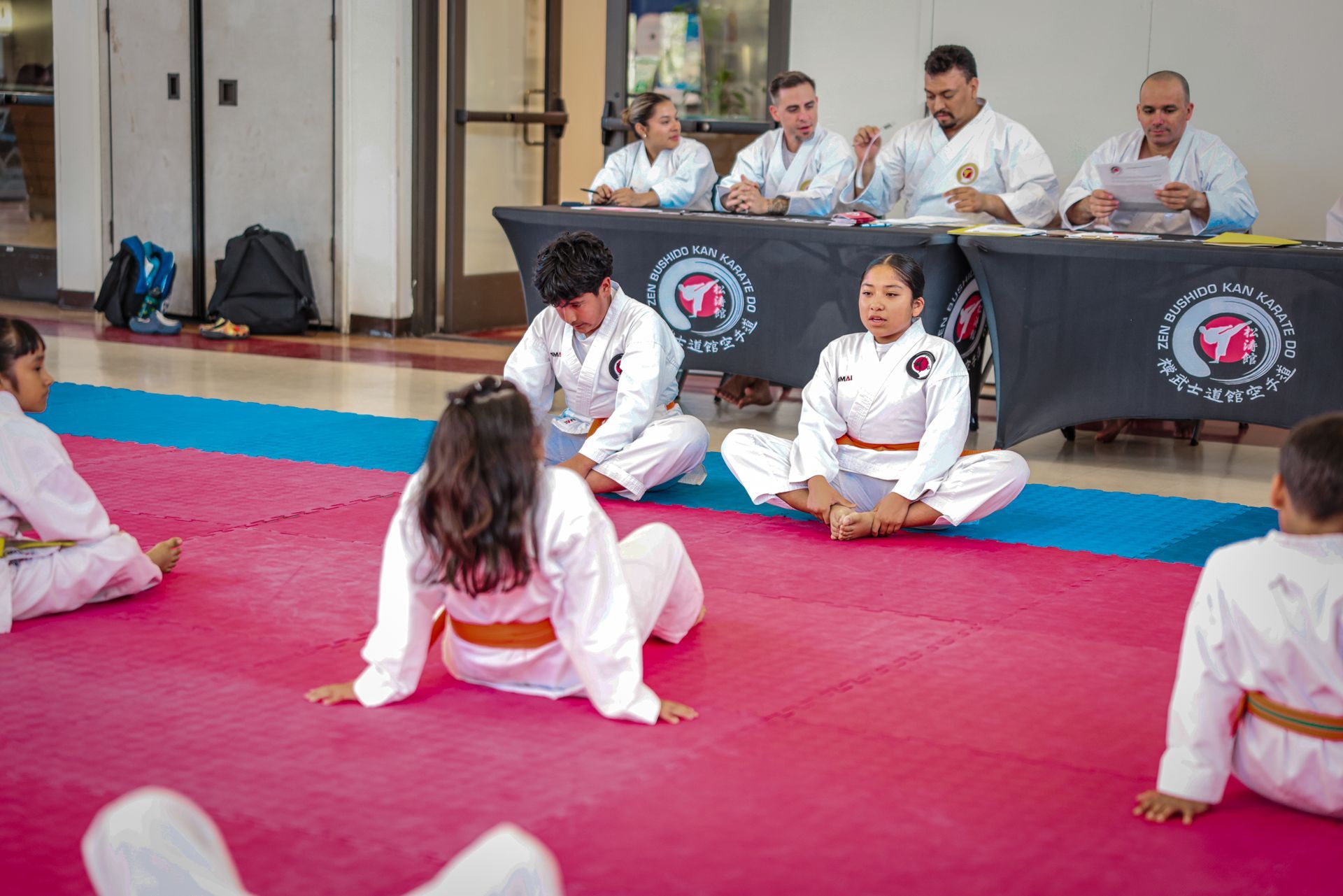 Students in white karate uniforms sit on a pink mat for a test, while four judges watch from behind a table.
