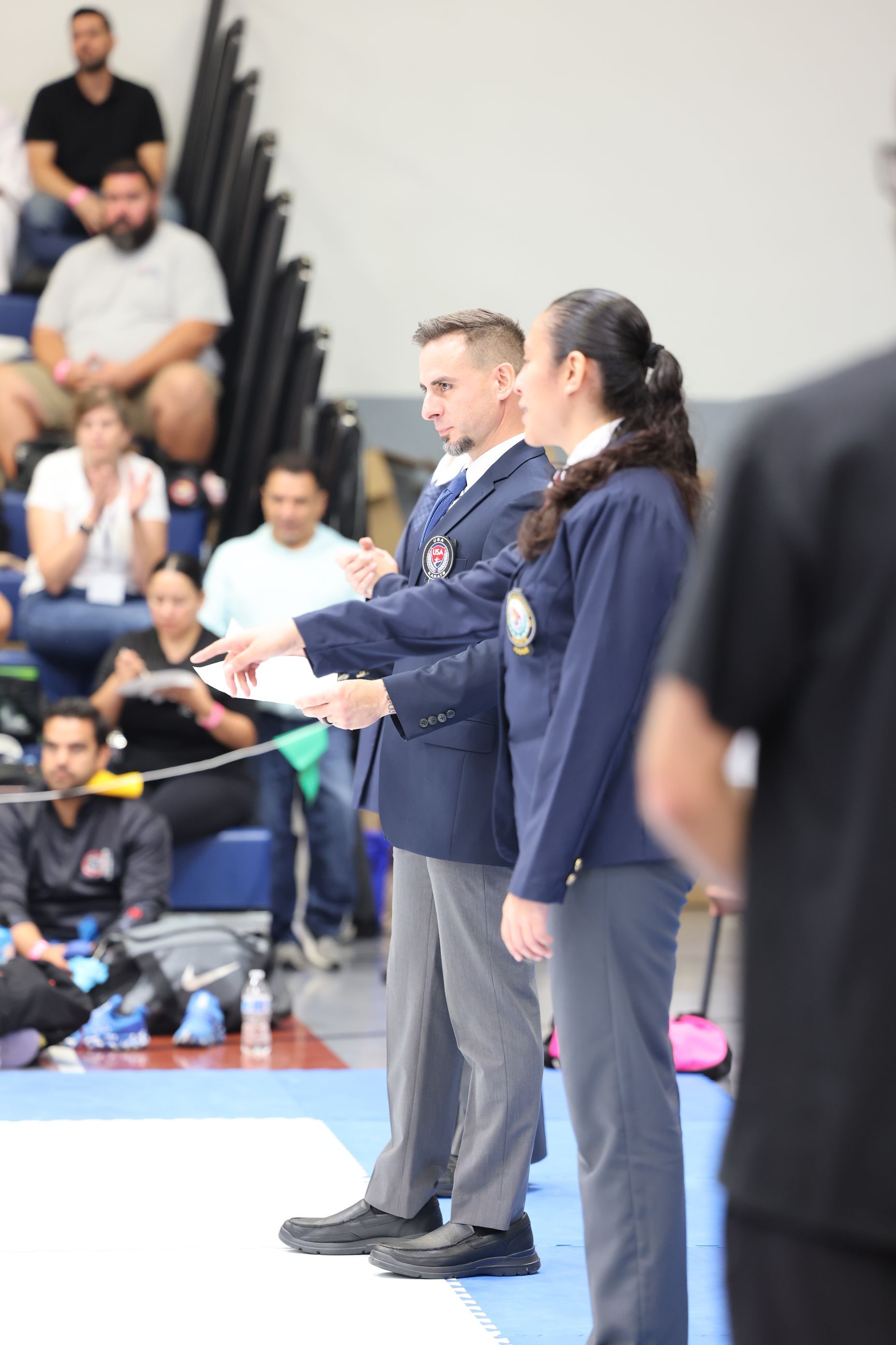 Karate referees gesturing at a competition, indoors, people in the background, spectators.