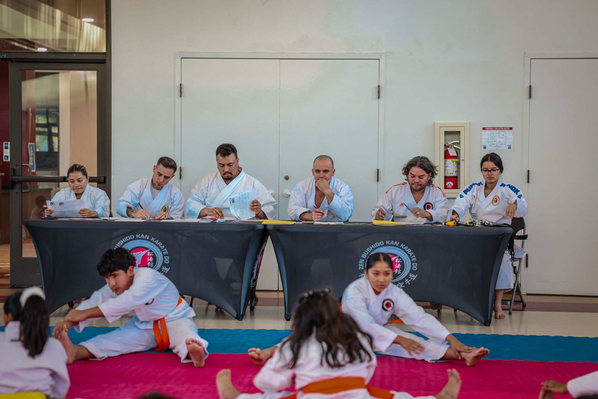 A group in karate uniforms stretches on a mat in front of a panel of judges seated at tables during a martial arts event.