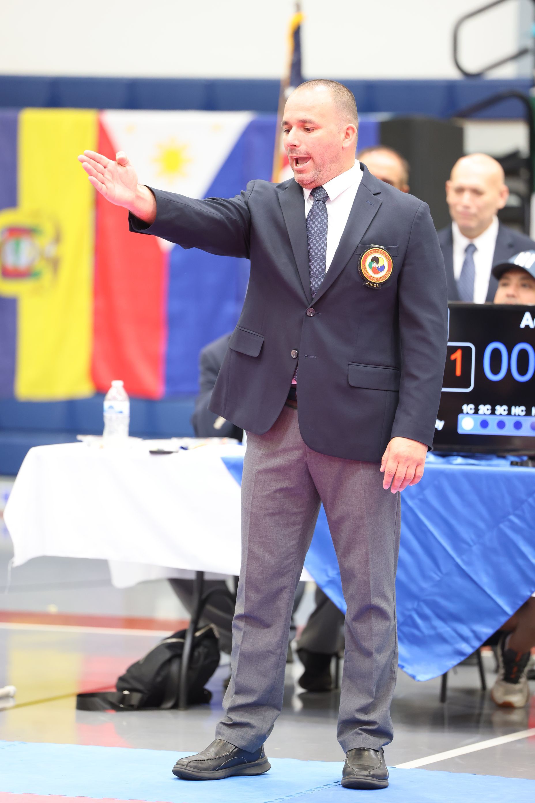 Referee in a dark blazer, gray pants, and tie gesturing with arm raised at a competition.