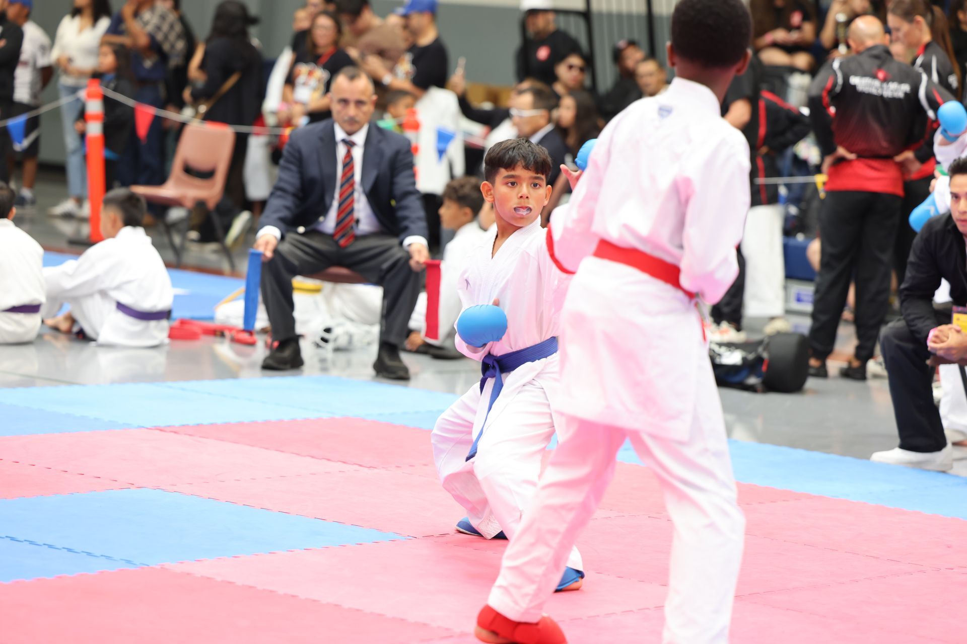 Two young karate competitors in white gis face off in a dojo. Referee watches.