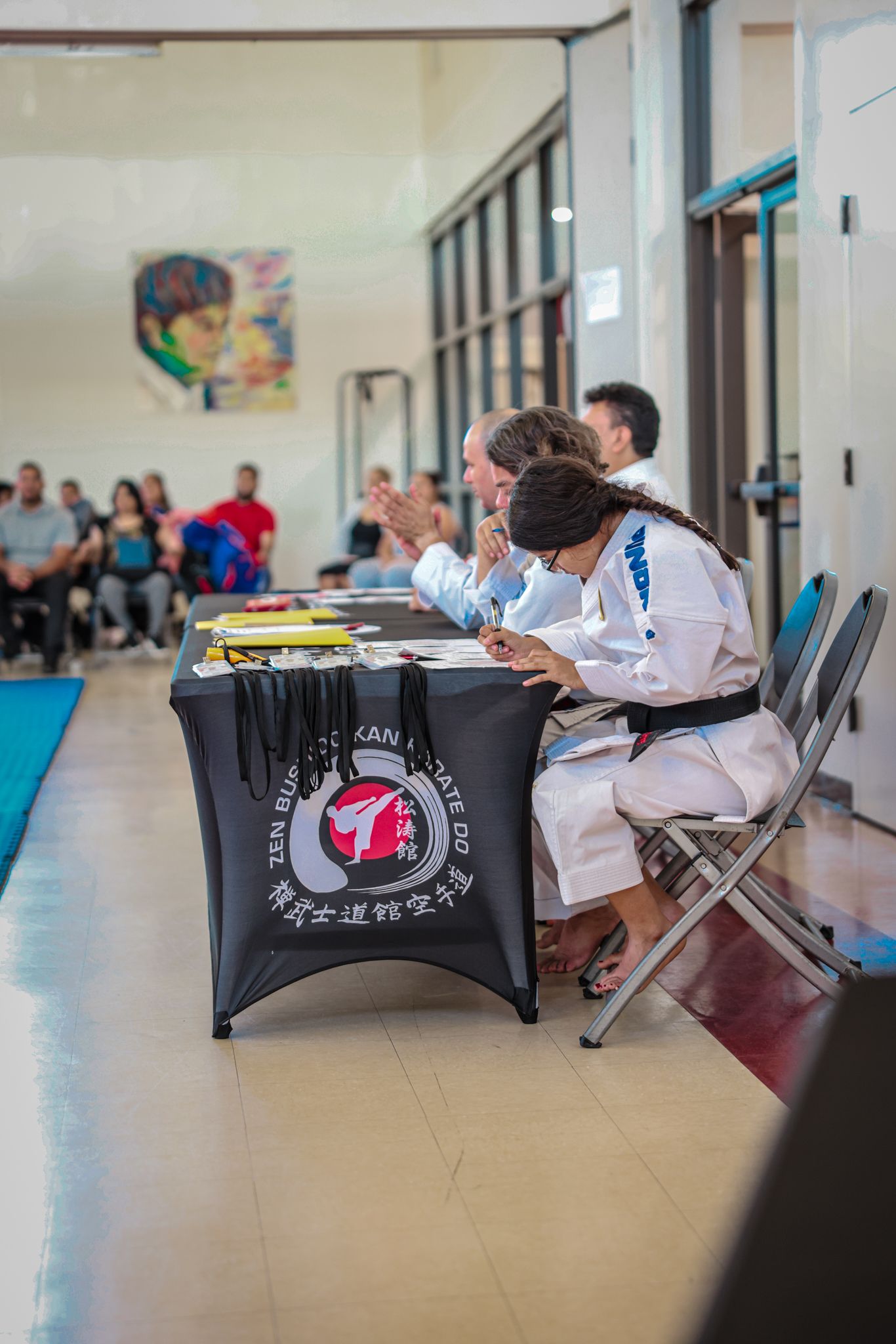 A row of judges in karate uniforms sits behind a table, reviewing documents during an indoor martial arts competition.