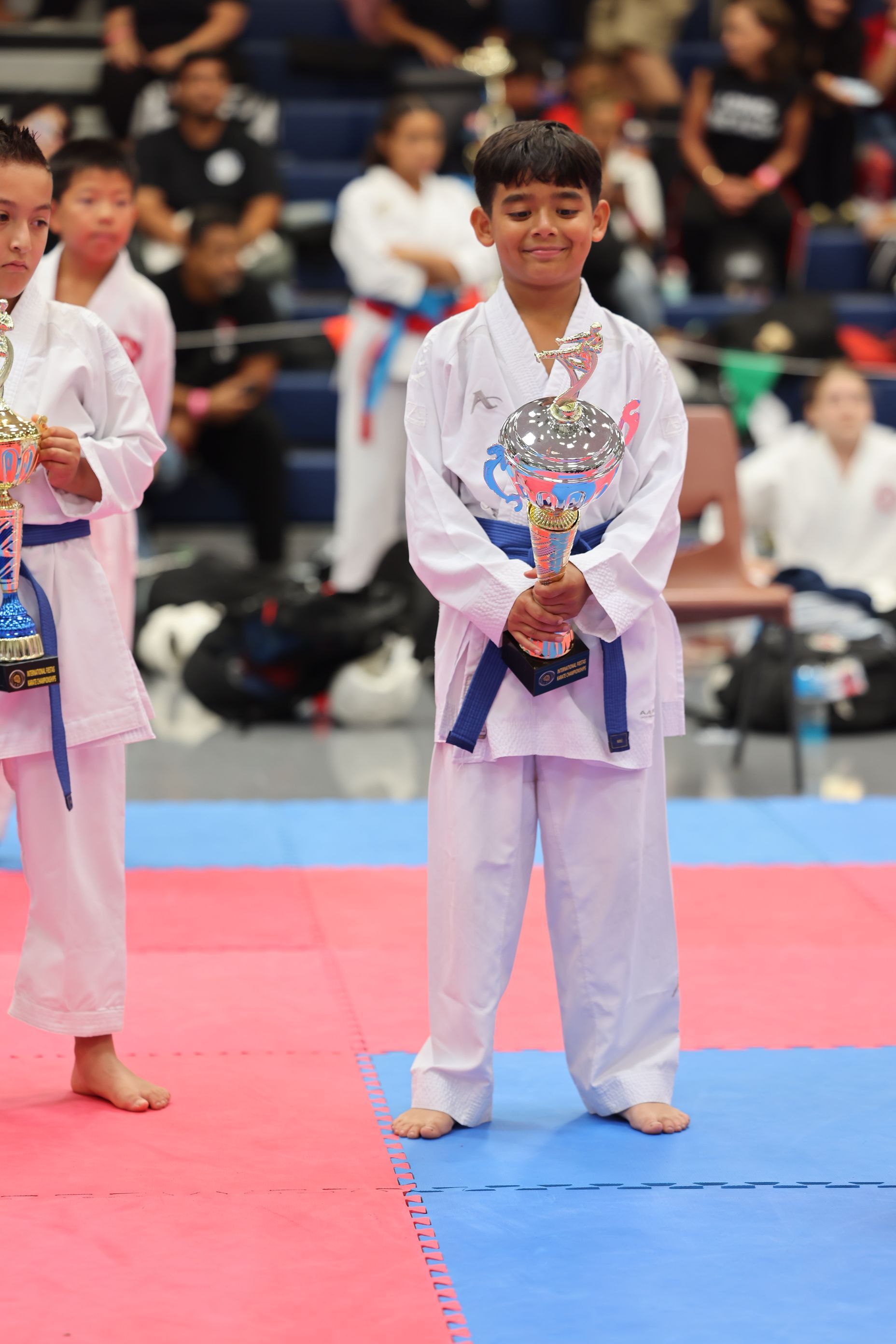 Boy in white karate uniform holding trophy, standing on mat at tournament.