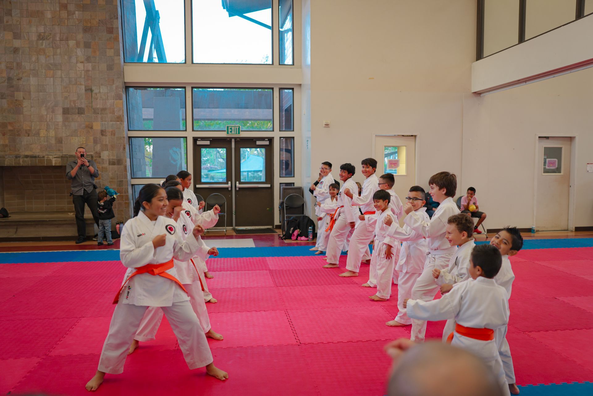 A group of students in karate uniforms with colored belts practicing martial arts moves on a pink and blue gym mat.