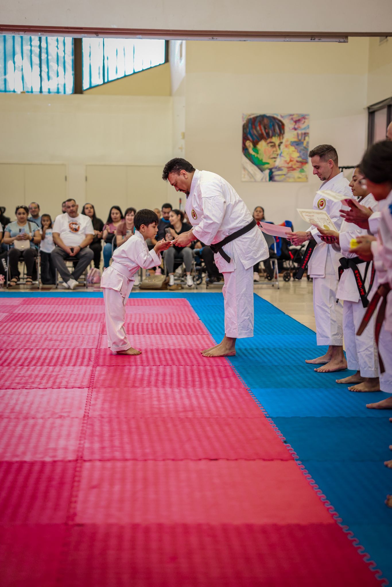 A child in a white karate uniform bows to an instructor in a black belt during a ceremony on a red and blue mat.