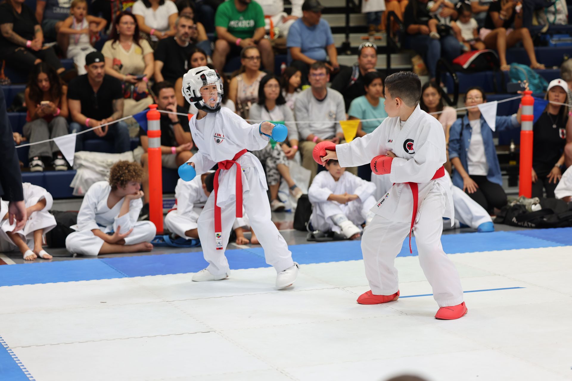Two young karate athletes in white uniforms with red belts sparring in a competition.