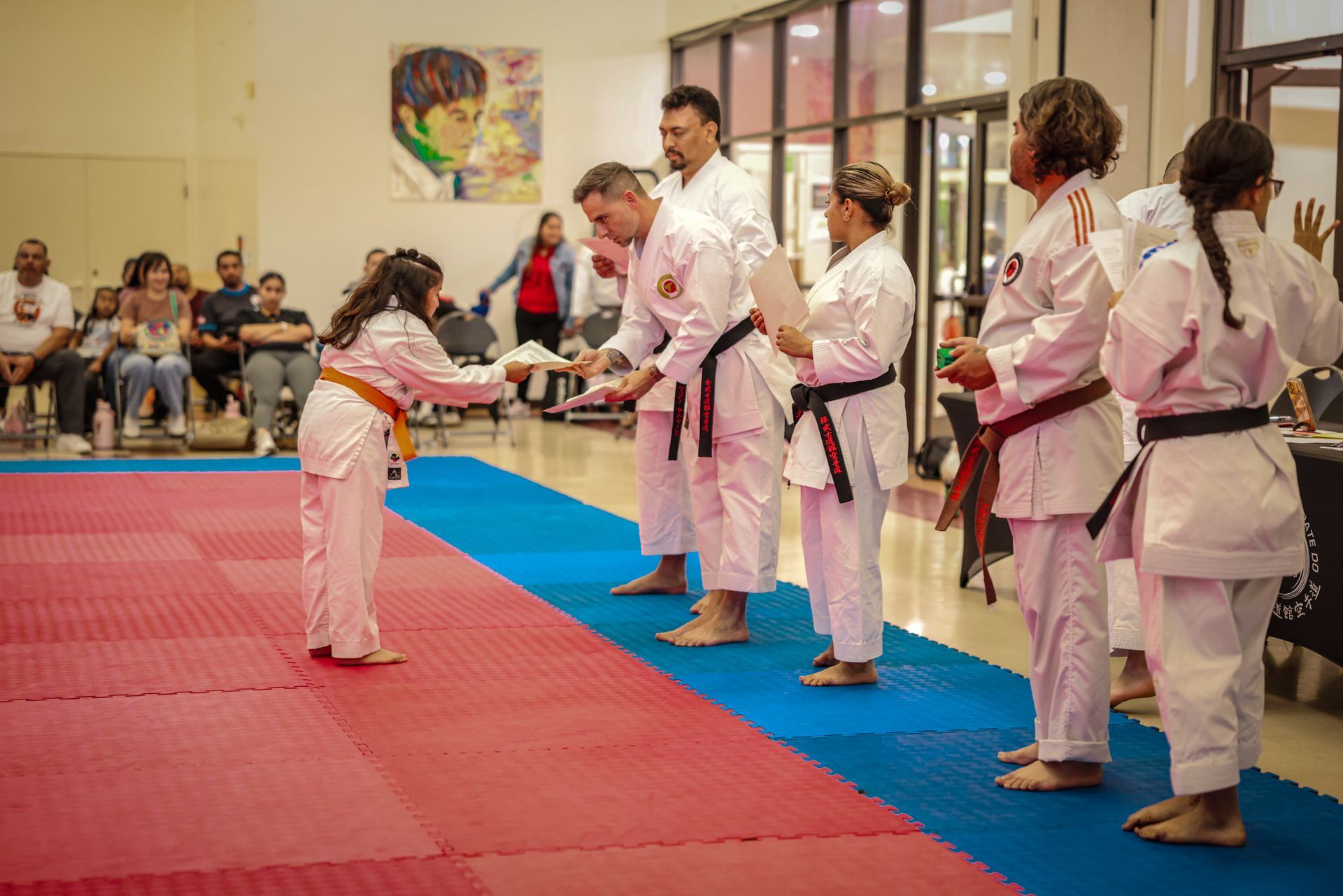 A martial arts student bows to an instructor on a red and blue mat, while other practitioners stand in a line nearby.