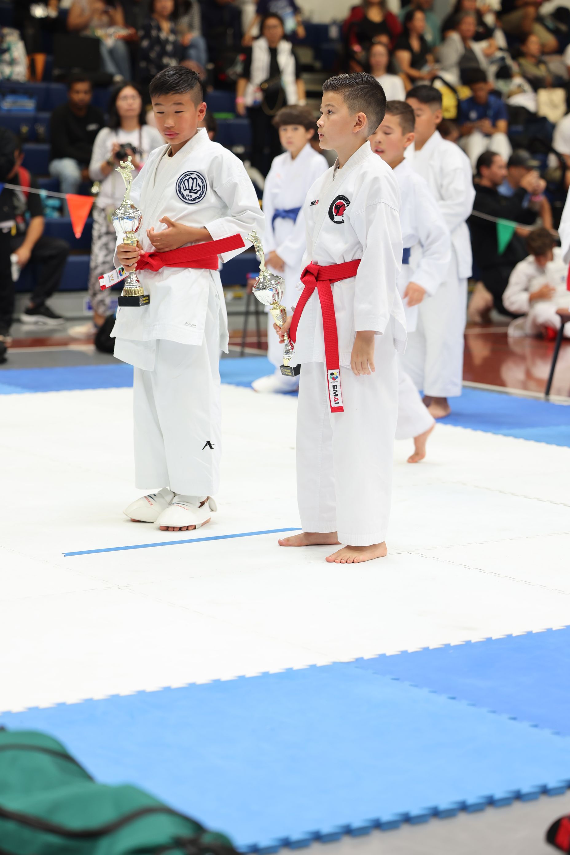 Boys in white karate uniforms with red belts, holding trophies on a blue mat.