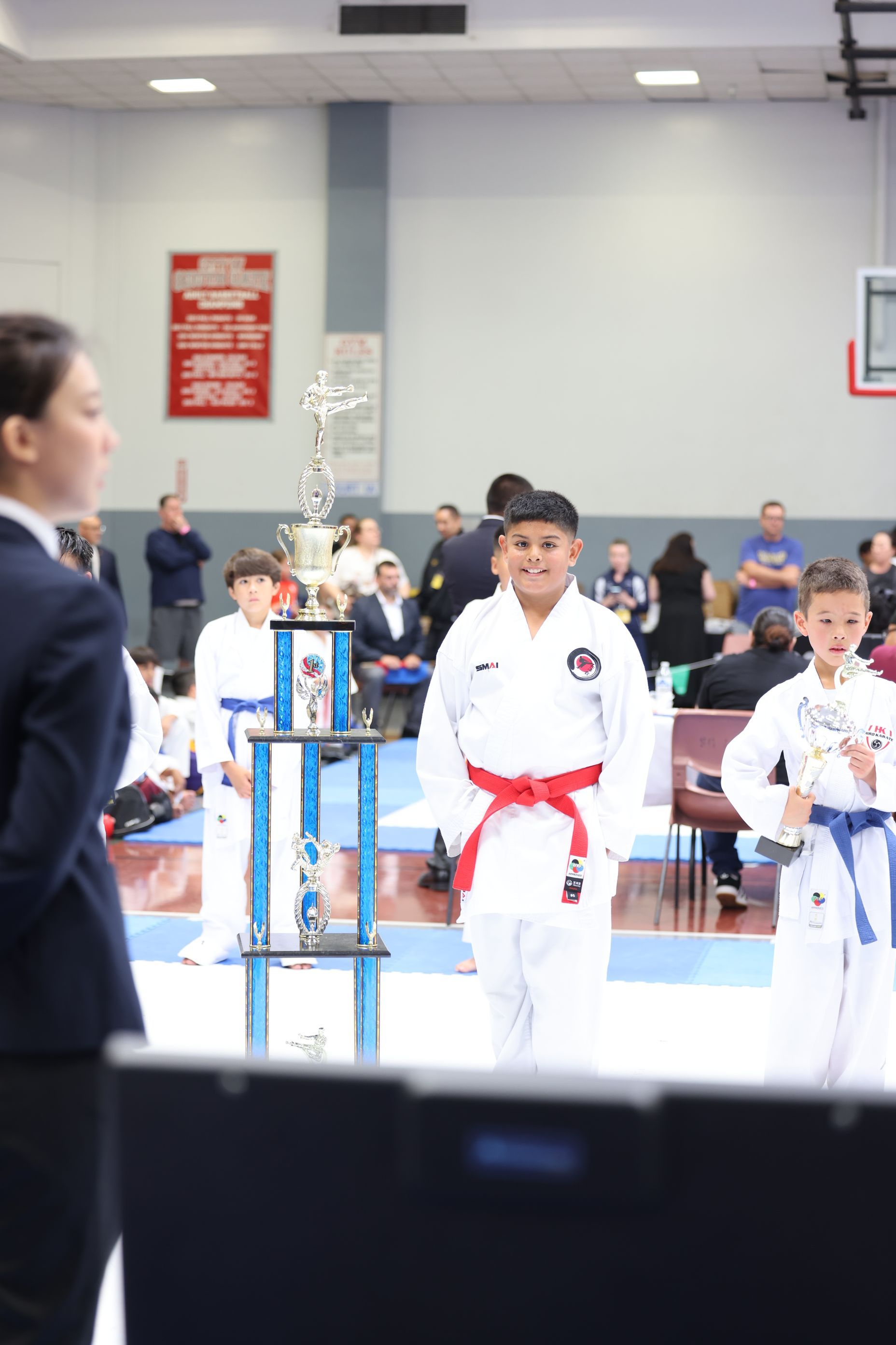 Boy in red belt smiling at karate tournament, trophy on stand. Other kids in the background.
