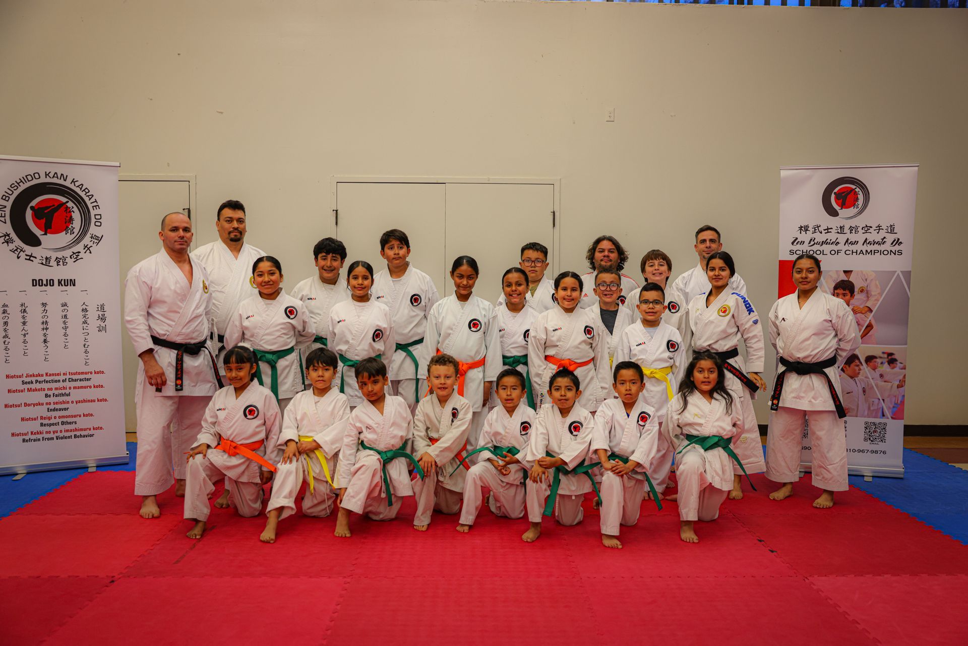 A group of martial artists in white uniforms with varying belt colors pose on a red mat in a room with two display banners.