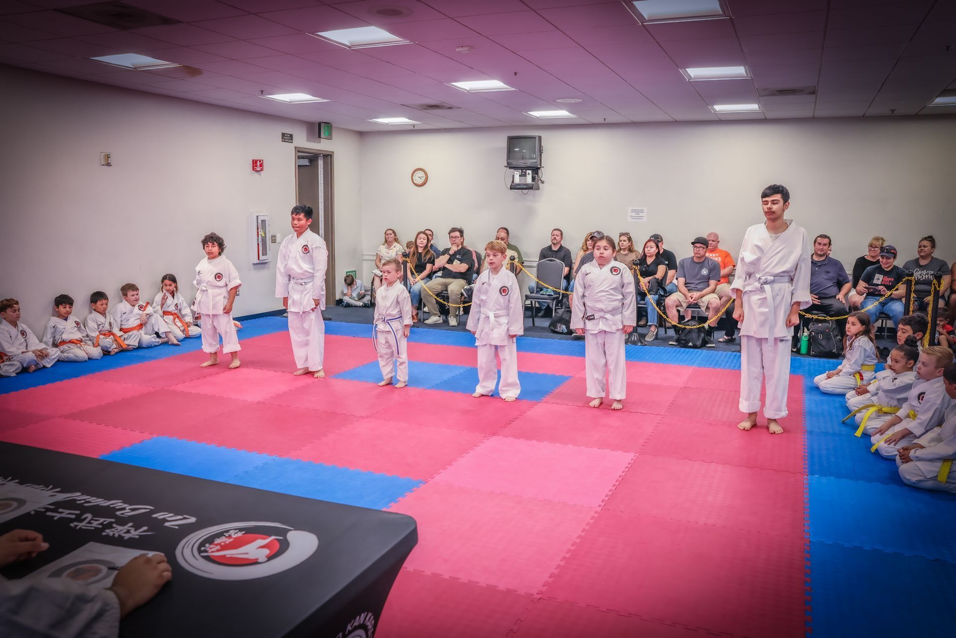 Children in martial arts uniforms stand on pink and blue floor mats in a training studio with an audience watching.
