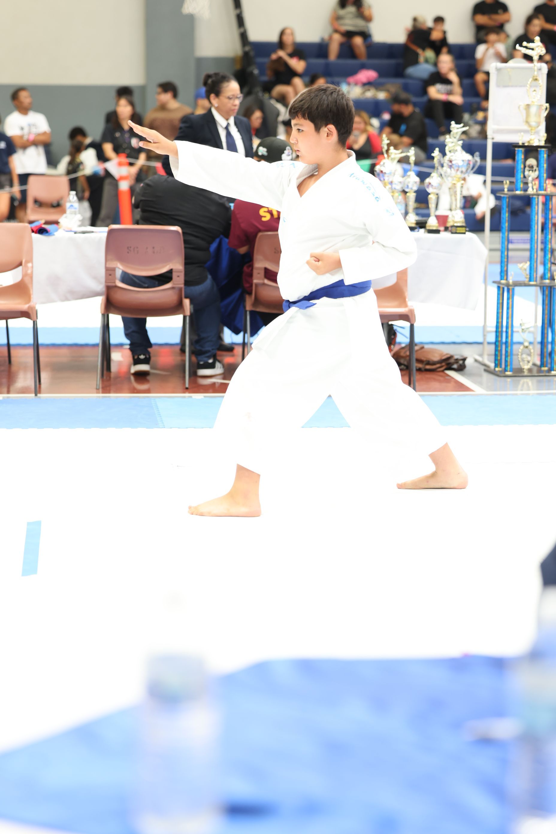 A young karateka in a white uniform with a blue belt performs a karate stance on a competition floor.
