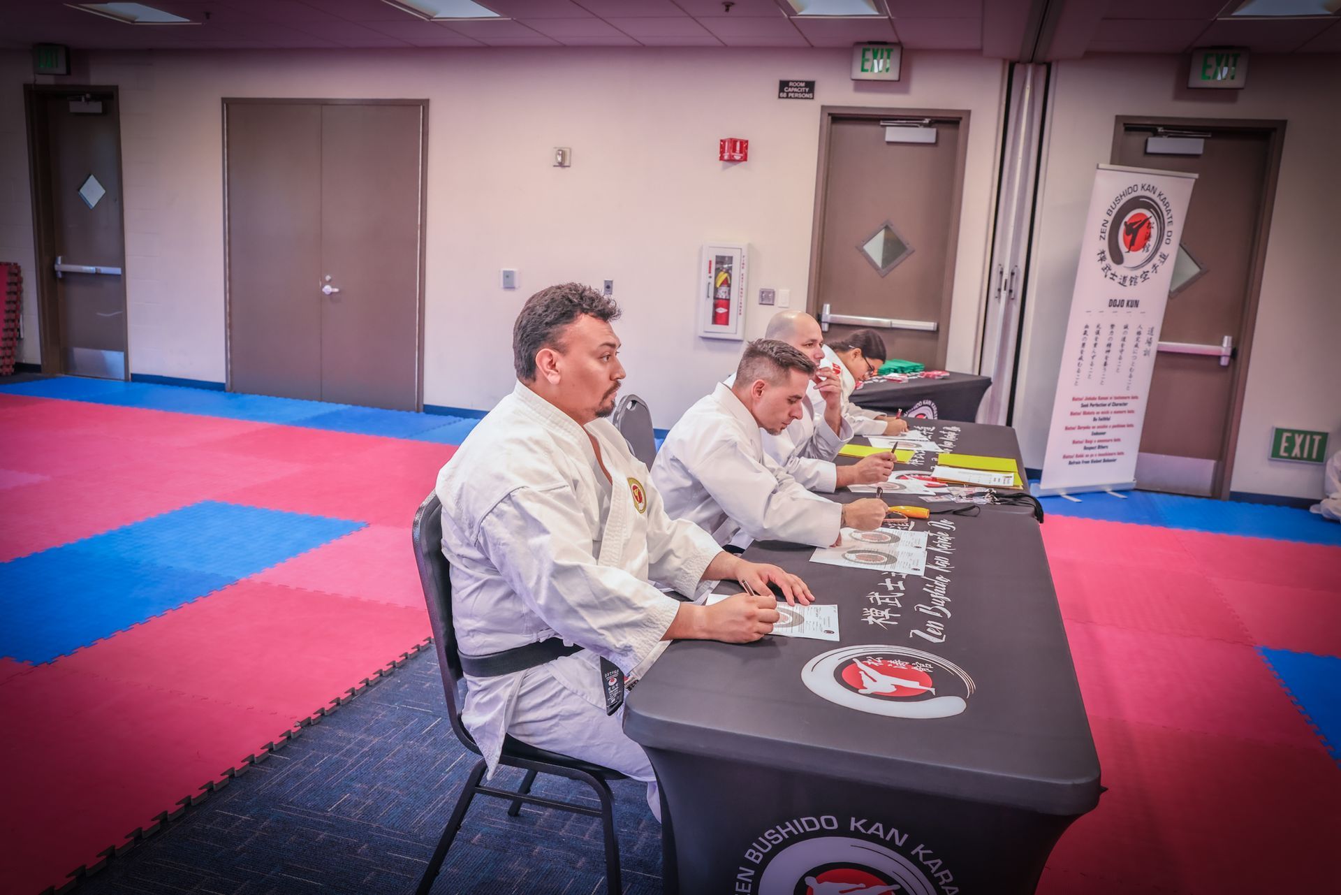 Three people in martial arts uniforms sit at a black table judging a competition in a room with blue and red floor mats.