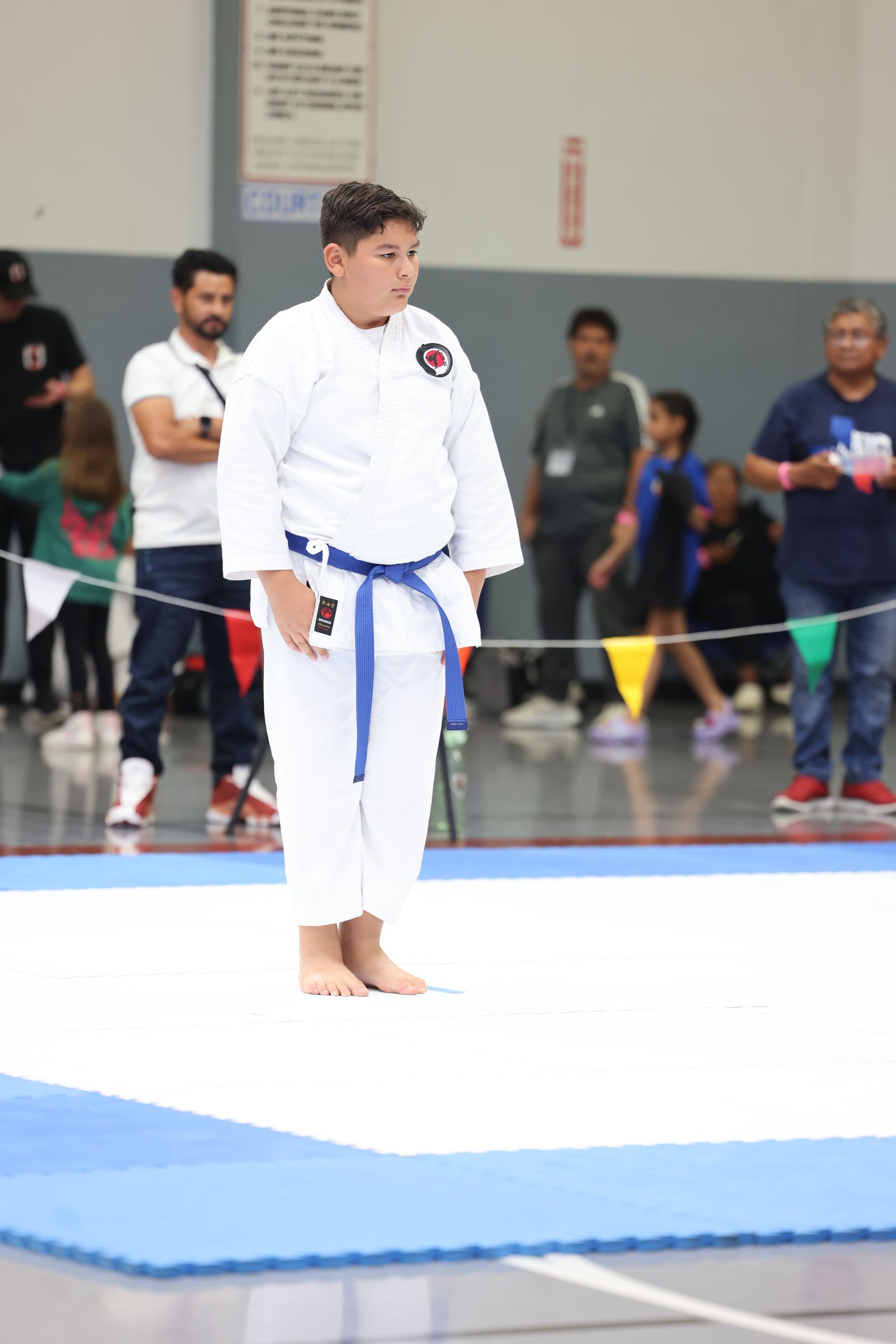 Boy in blue belt karate uniform on a mat, awaiting competition; indoors, other people watching.