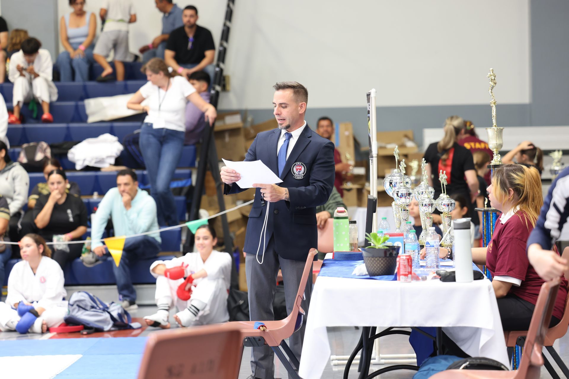 Referee at karate tournament reads notes. Spectators watch from bleachers.