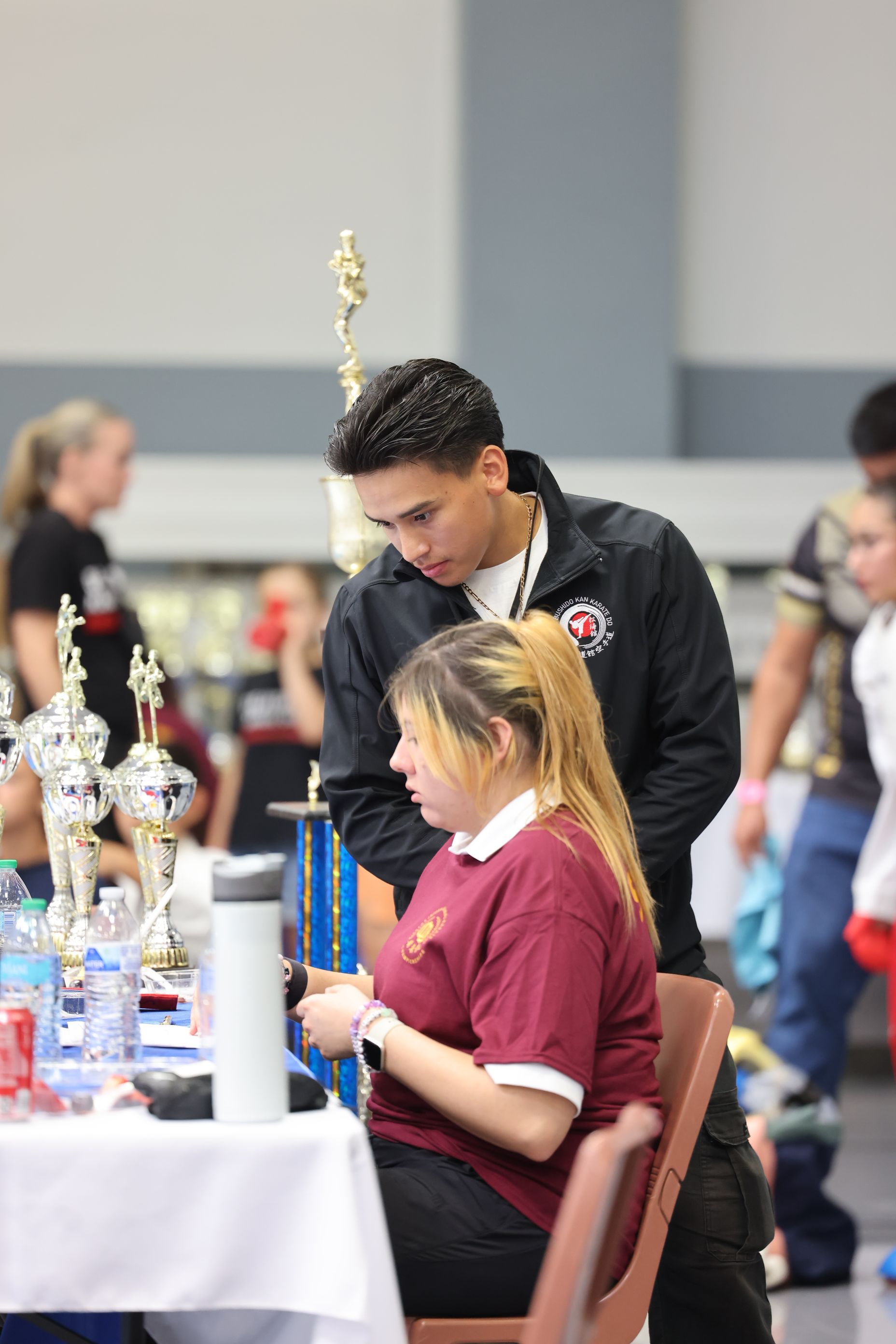 Man in black jacket leans over a seated girl in a maroon shirt, in a competition setting.