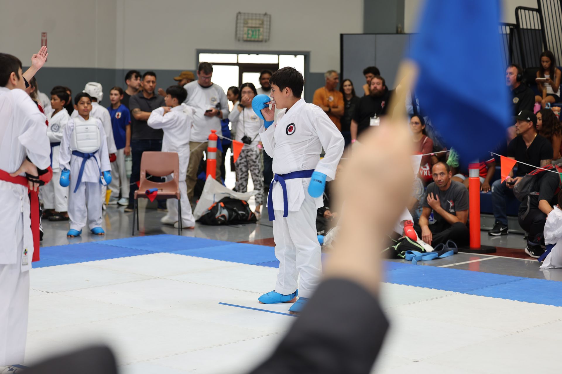 Karate match: boy in blue uniform bows as judge raises blue flag. Other competitors and spectators watch.
