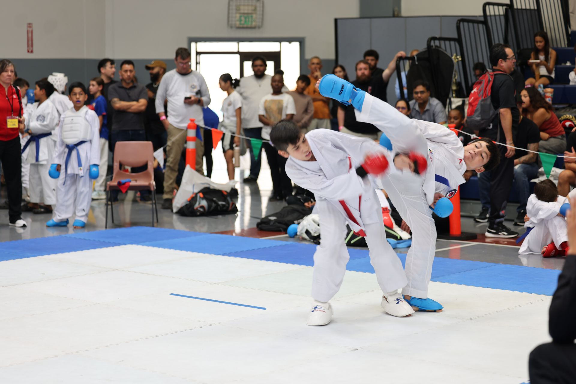 Two karate competitors in white uniforms, one in mid-air performing a kick. Spectators watch.