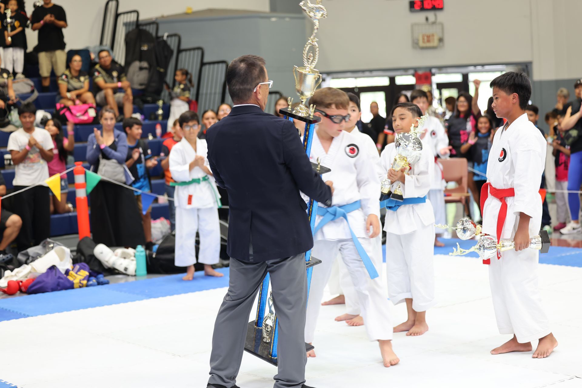 Karate tournament award ceremony; children in white gis hold trophies, adults clap.