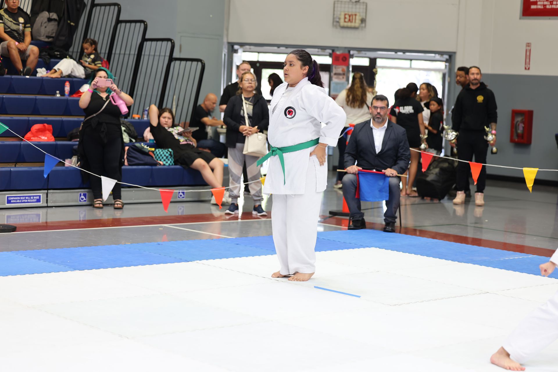 Female karateka in white uniform with green belt, standing on a mat, hands on hips, at a tournament.