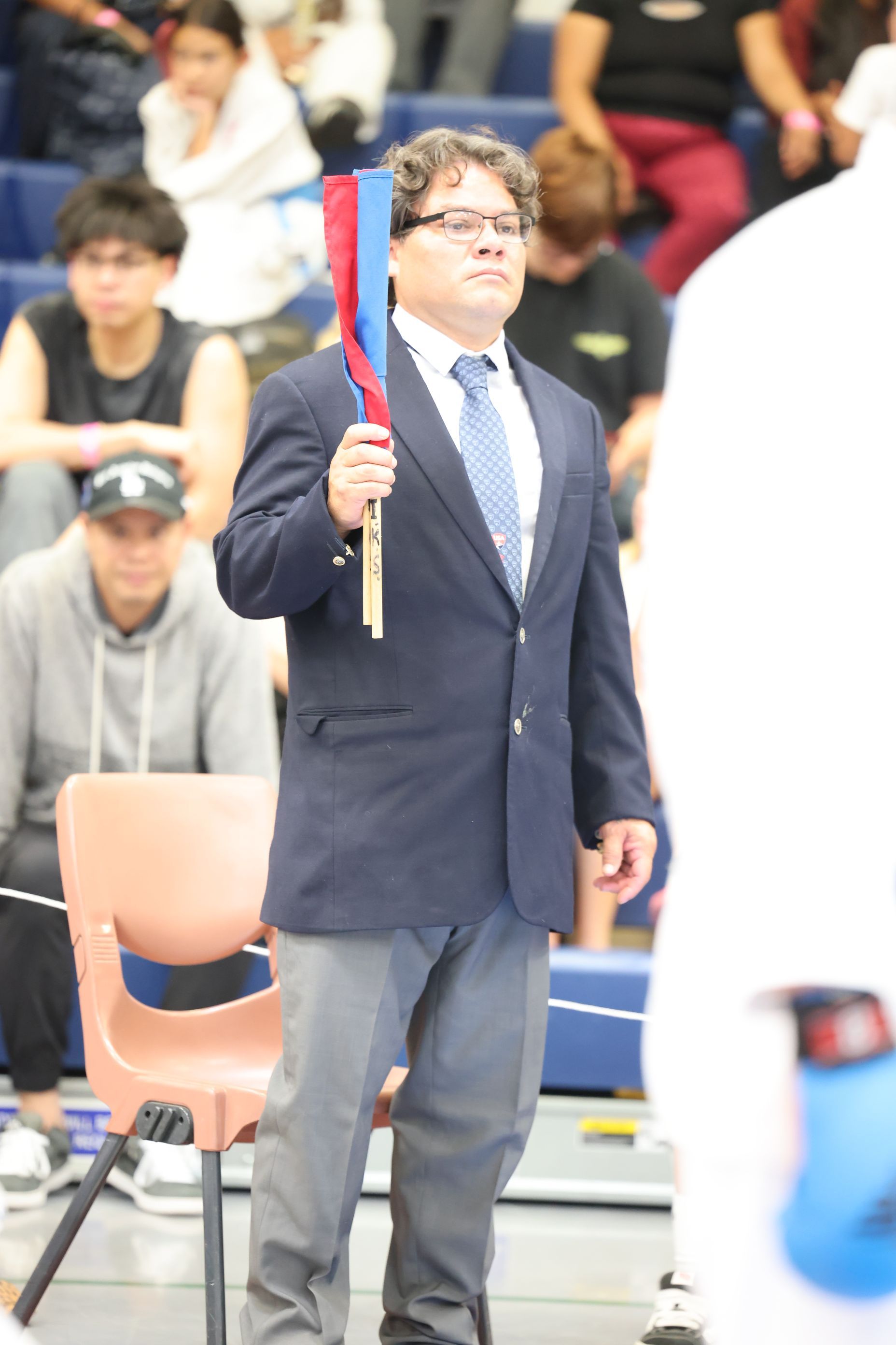 Man in blazer and tie holding red and blue flag at event, spectators in background.