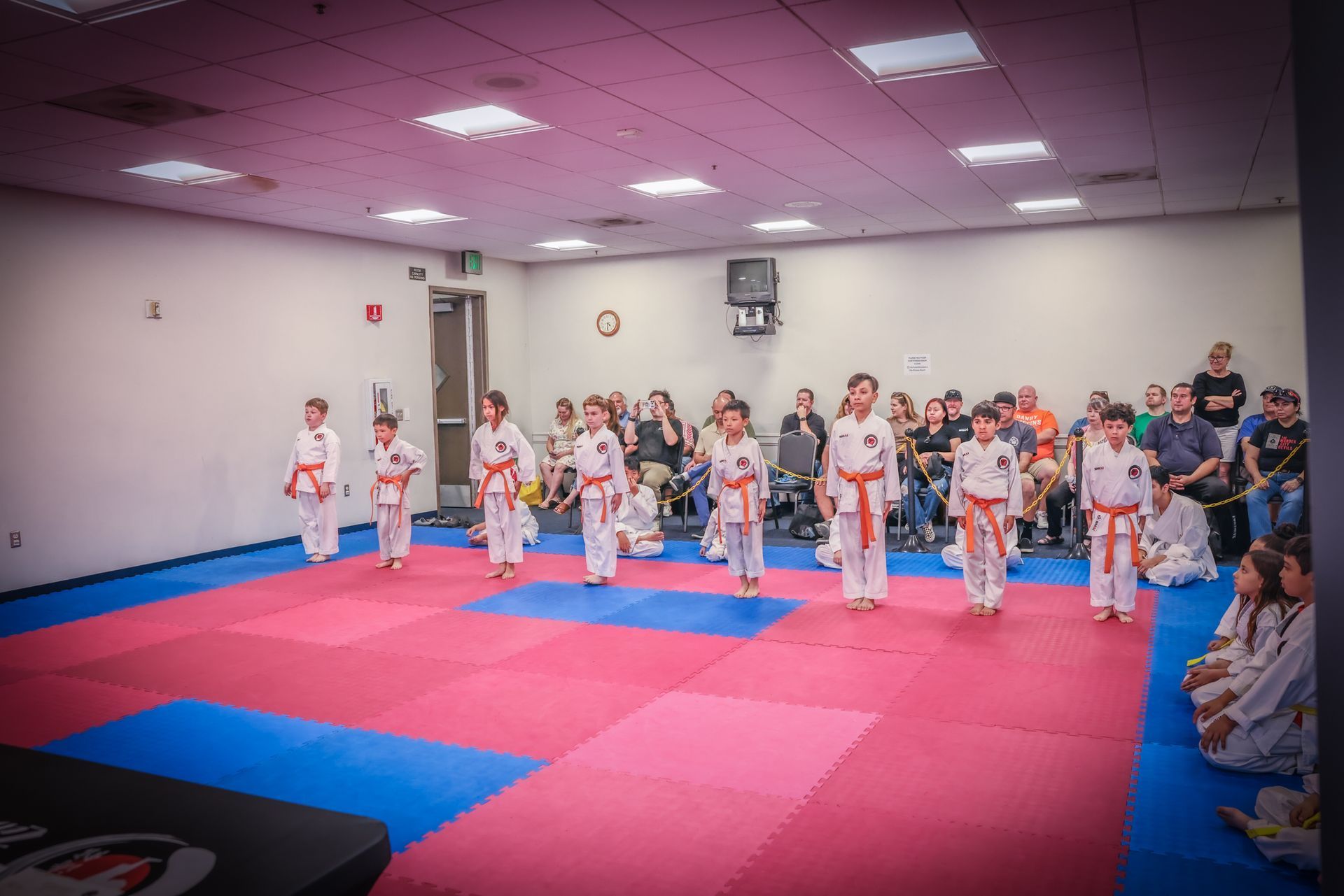 Students in white martial arts uniforms with orange belts stand in a line on a red and blue padded floor in a studio.