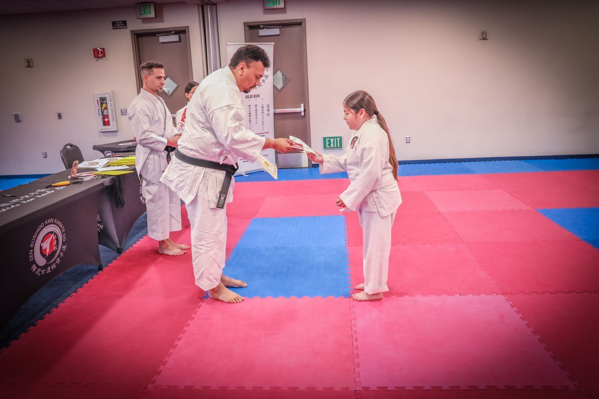 A martial arts instructor in a white gi hands a certificate to a student in a white gi on a red and blue mat.