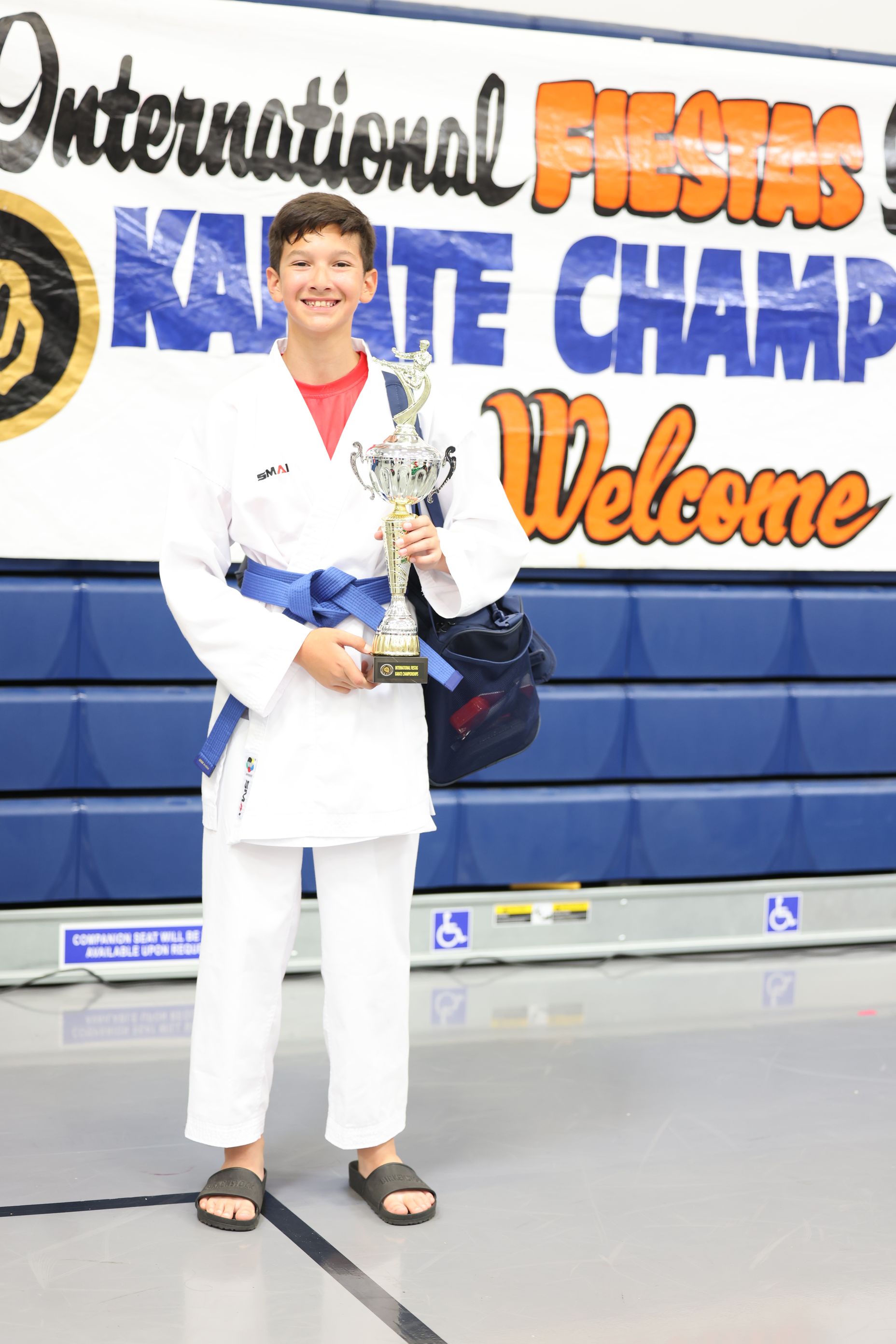 Young karate champion in a blue belt, holding a trophy, standing in front of a banner.