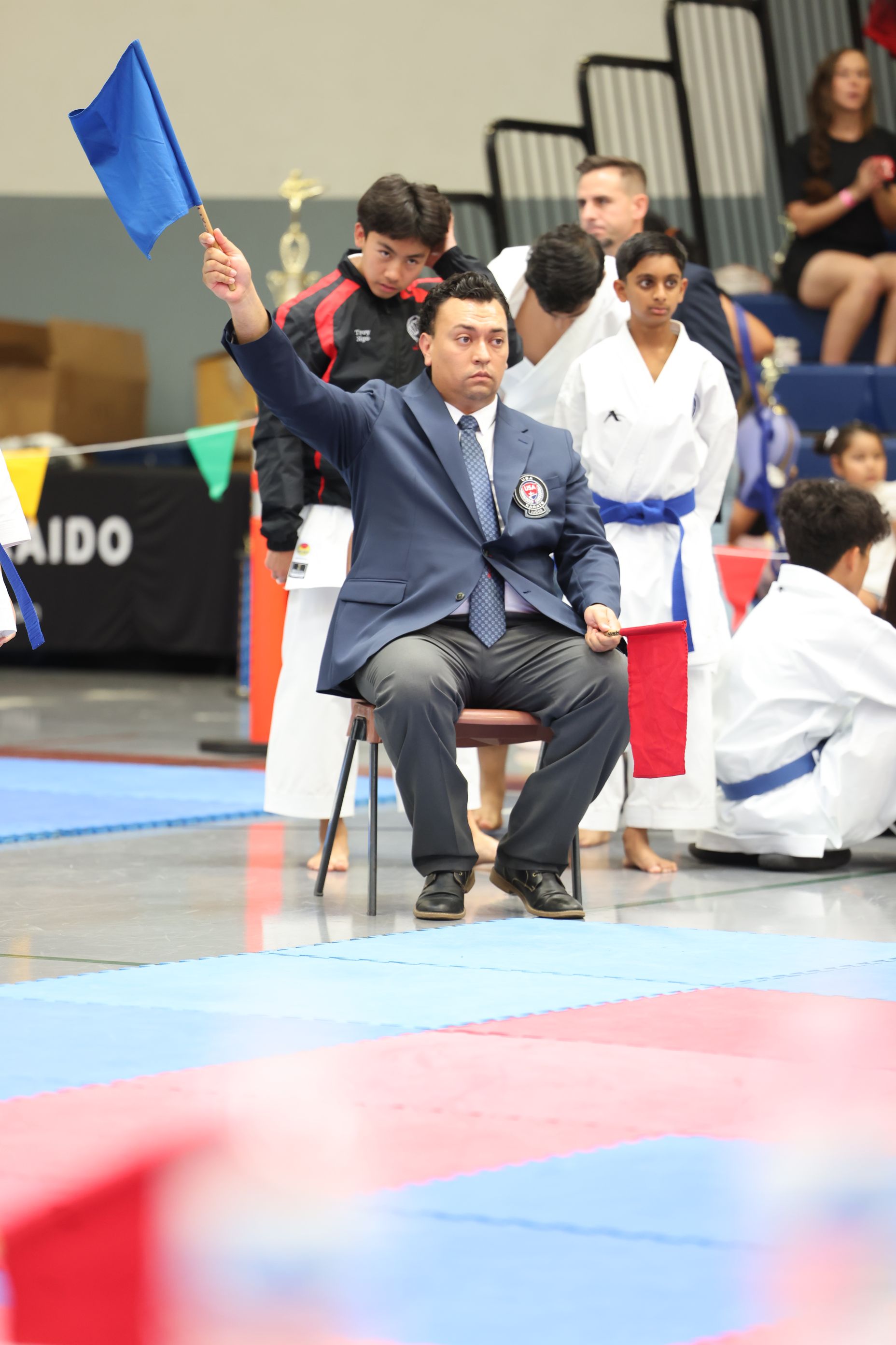 Karate judge in blue blazer raises blue flag, seated, in a tournament.