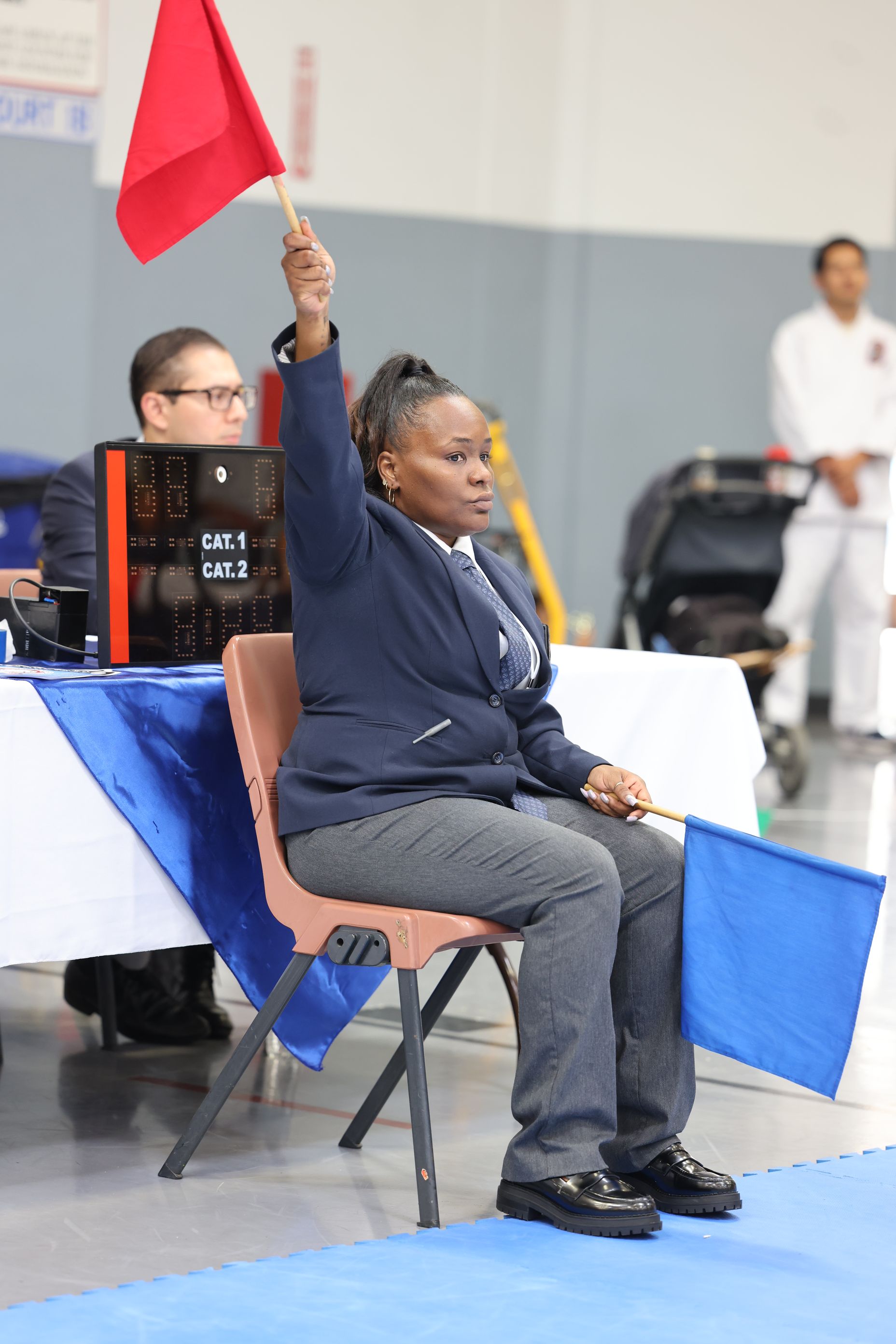 Woman in blazer raises red flag, seated at karate tournament judging table.