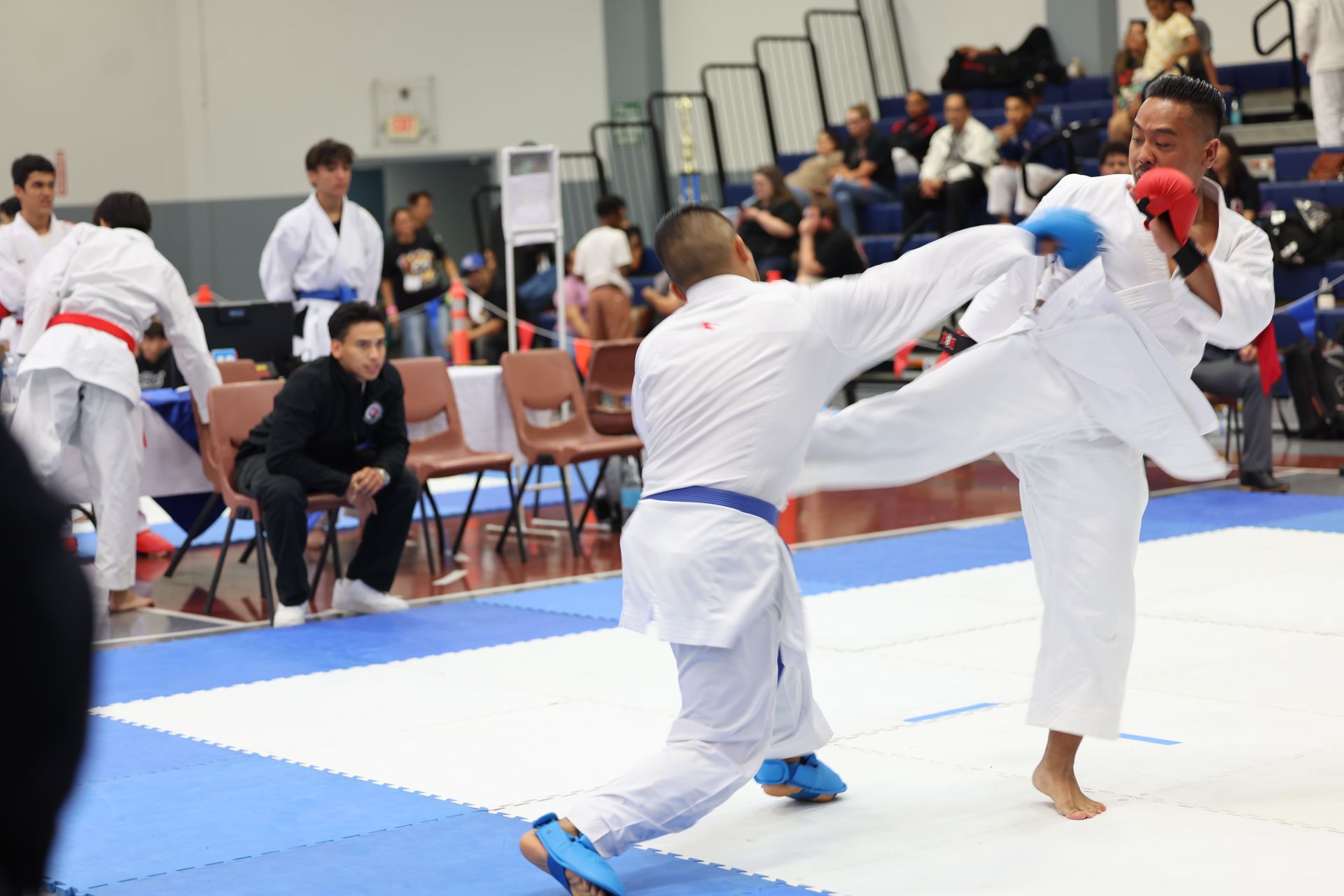 Karate sparring match. Two men in white uniforms with blue and red protective gear, one kicking. Indoor arena.