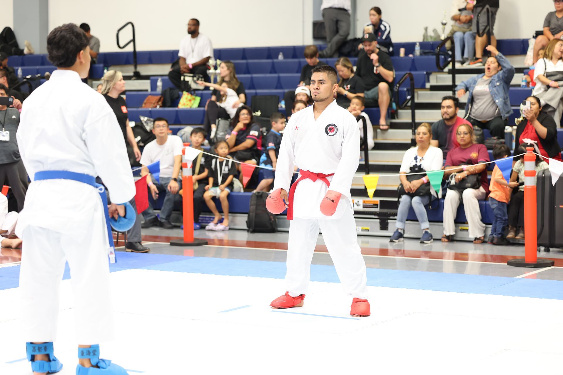 Two karate competitors face off on a blue mat, wearing white gis with blue and red belts, watched by an audience.