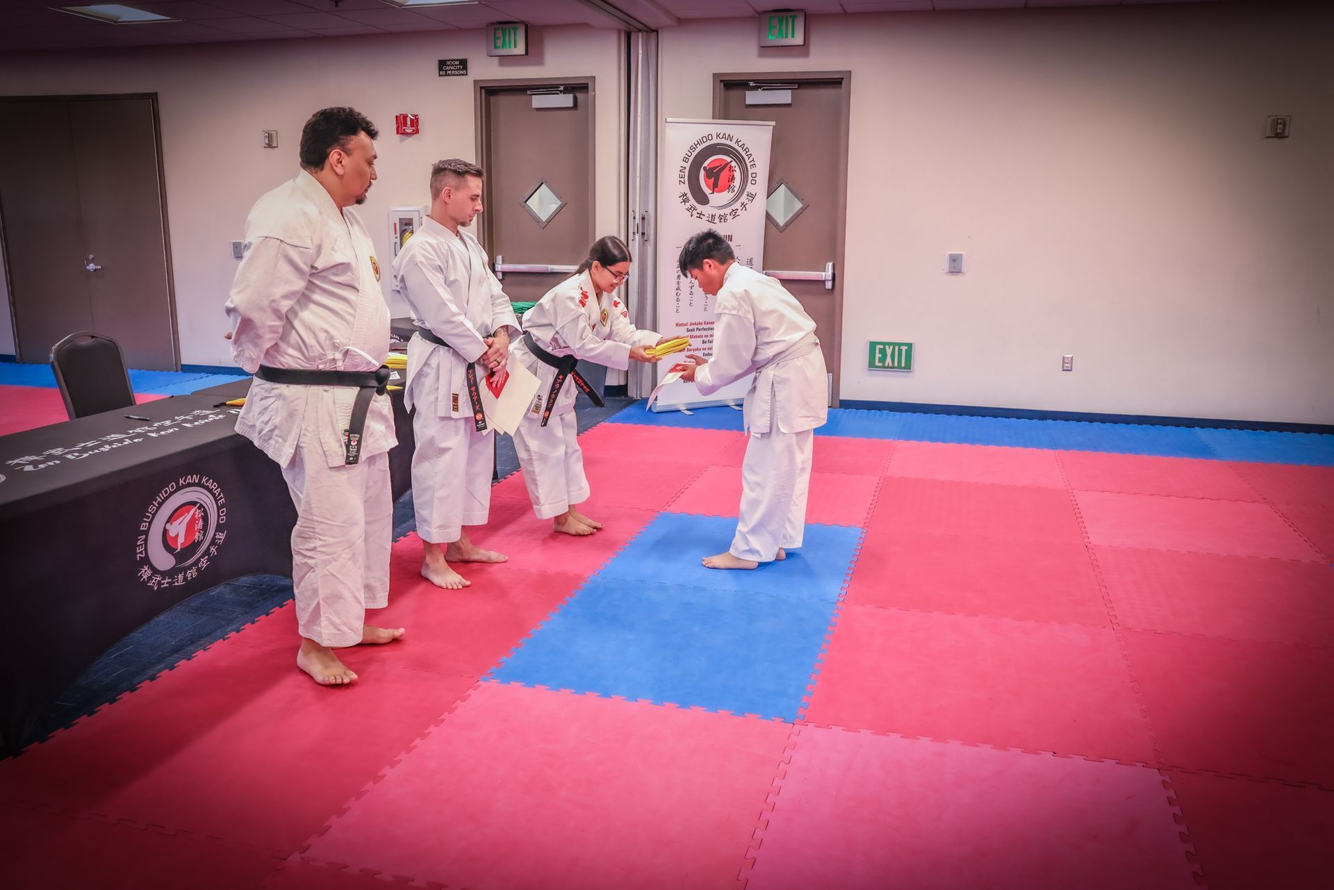 A student bows to an instructor to receive a yellow belt on a red and blue mat inside a martial arts studio.