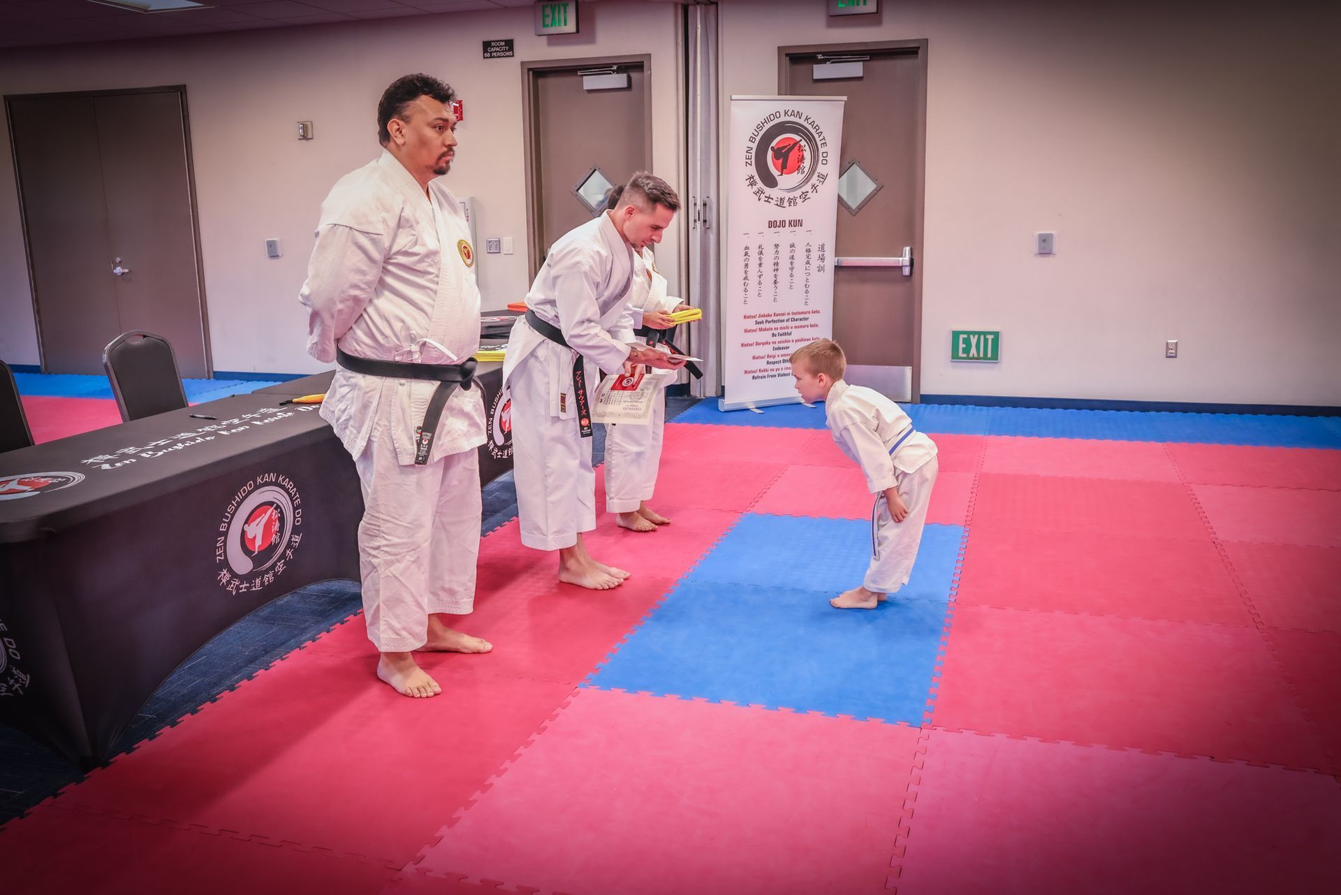 A child bows to an instructor on a blue and pink mat in a martial arts studio; a judge stands nearby to the left.