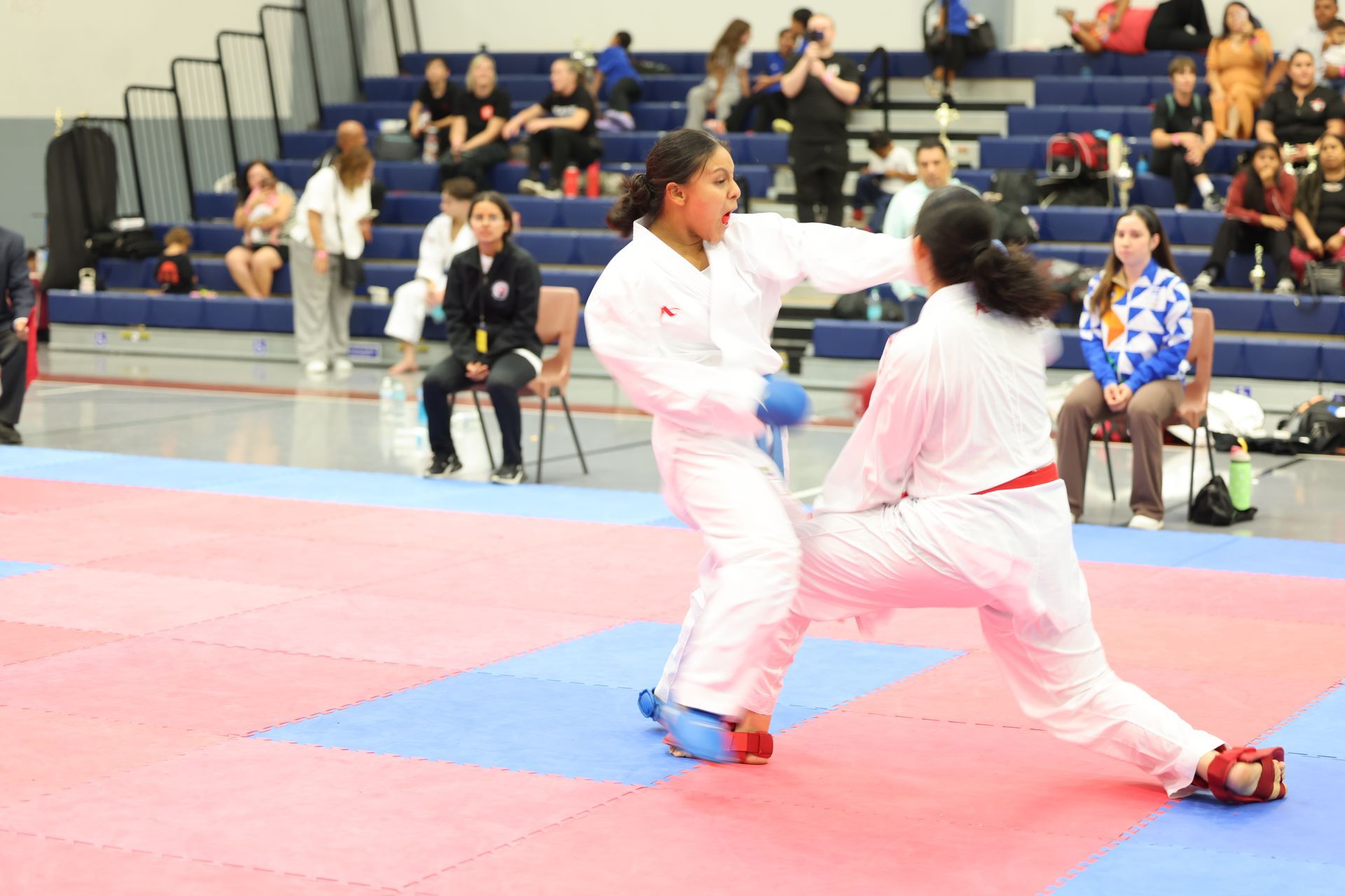 Two karate athletes in white uniforms compete on a mat; one throws a punch, the other is on the ground