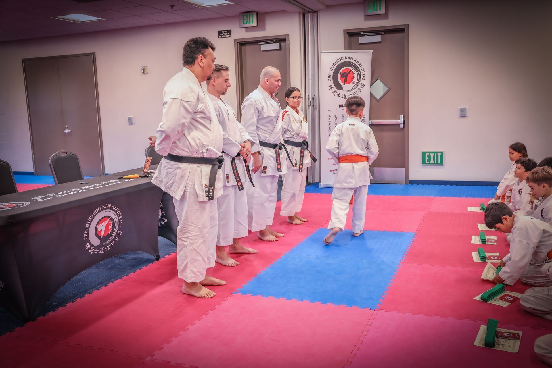 A line of martial artists in white uniforms and black belts stand facing a student in an orange belt in a gym setting.