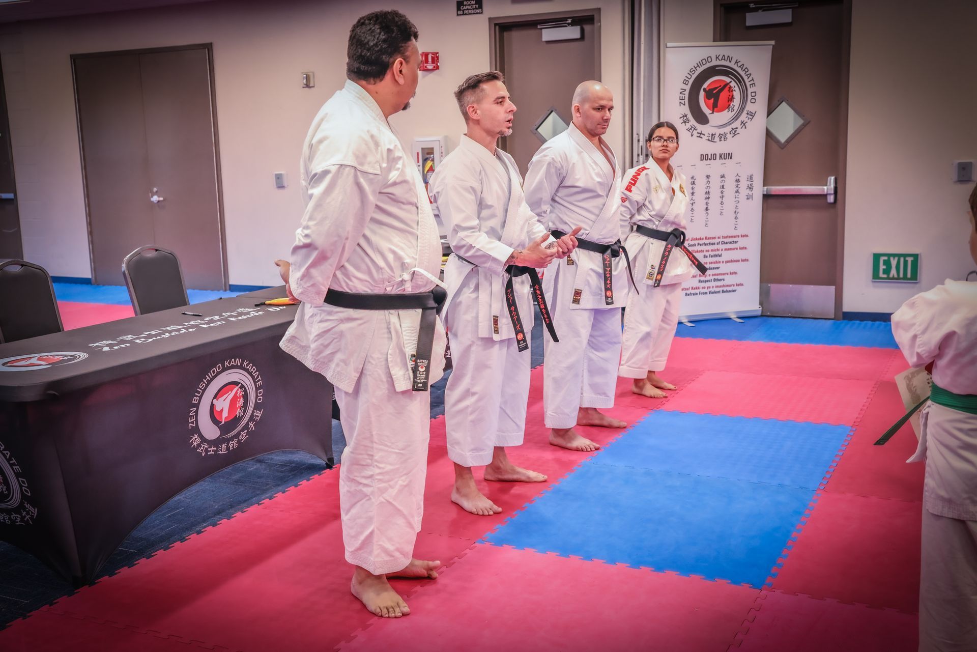 Four martial artists in white uniforms with black belts stand in a line on a training mat in front of a seminar banner.