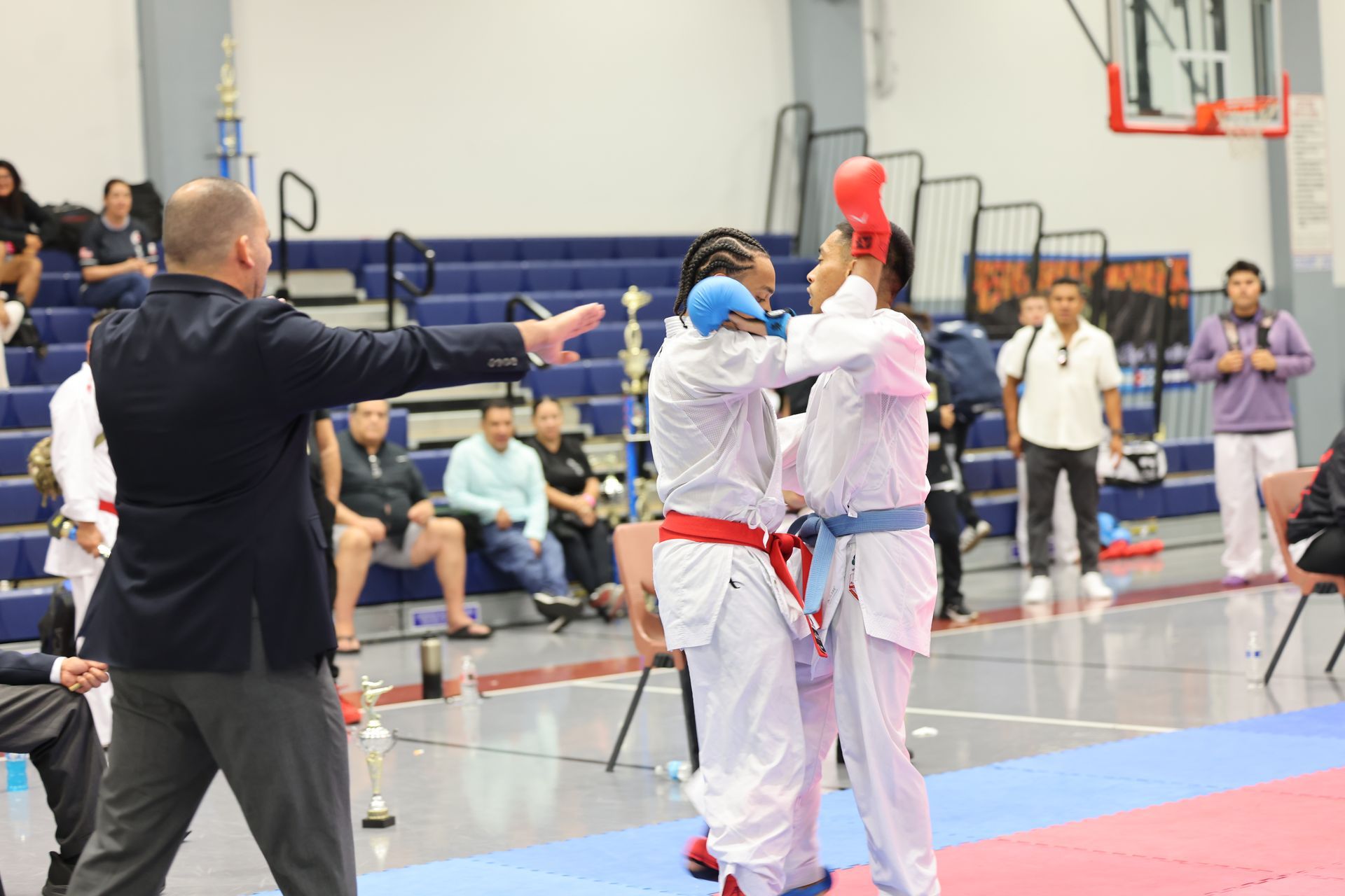 Karate match: Referee pointing; two competitors in white uniforms, blue and red protective gear, inside a gym.