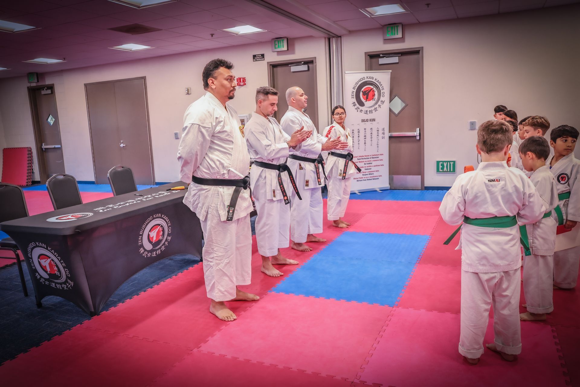 A group of karate students and instructors in white uniforms stand in rows on a red and blue training mat in a hall.