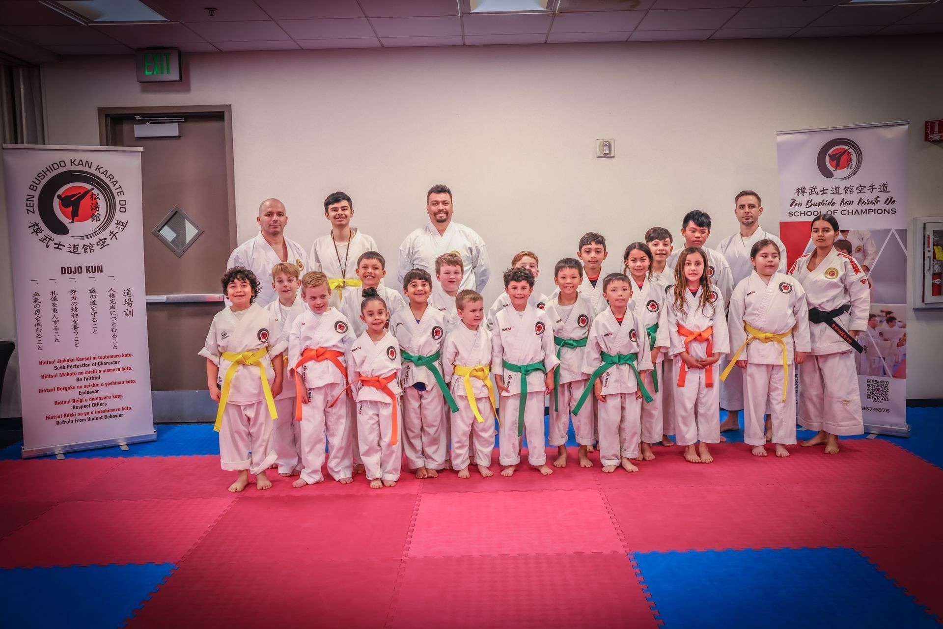 A group of karate students and instructors in white uniforms with colored belts posing in a training studio.